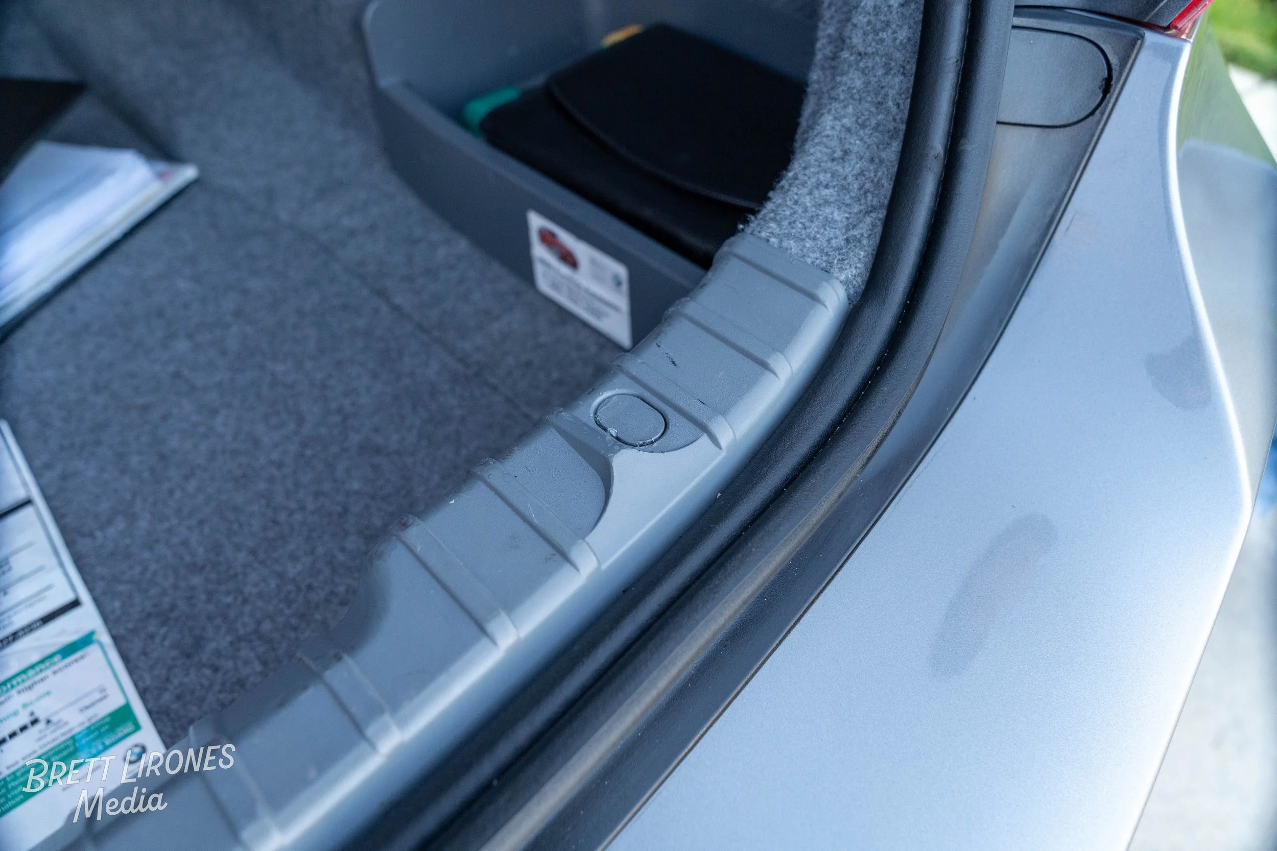 Close-up of a car trunk corner showing gray plastic trim, with a circular indentation and a black rubber seal. The trunk interior is carpeted gray with a black storage box and some papers visible inside.