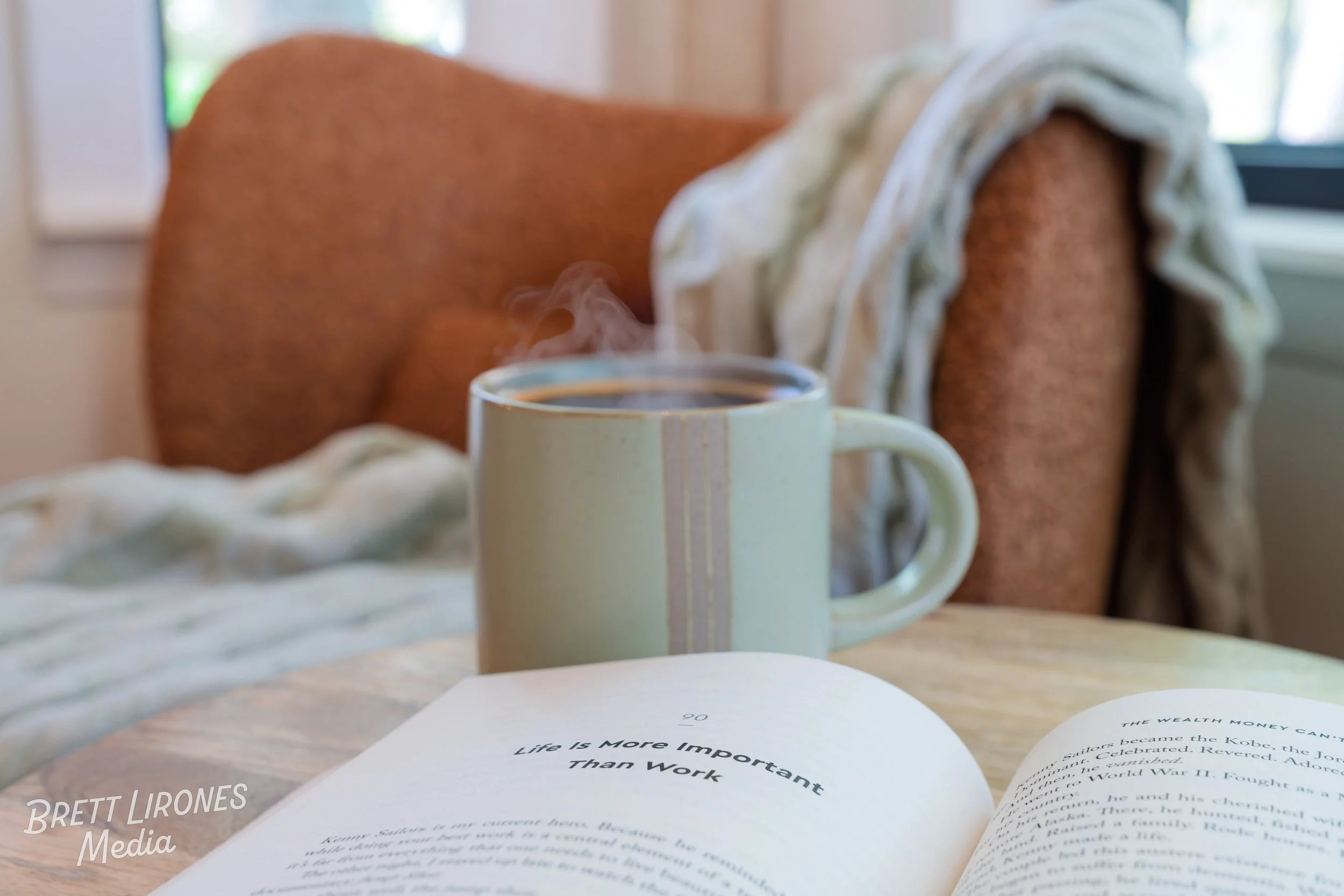 A steaming cup of coffee on a wooden table with an open book beside it. In the background, there's an upholstered chair with a blanket draped over it and a window with natural light coming through.