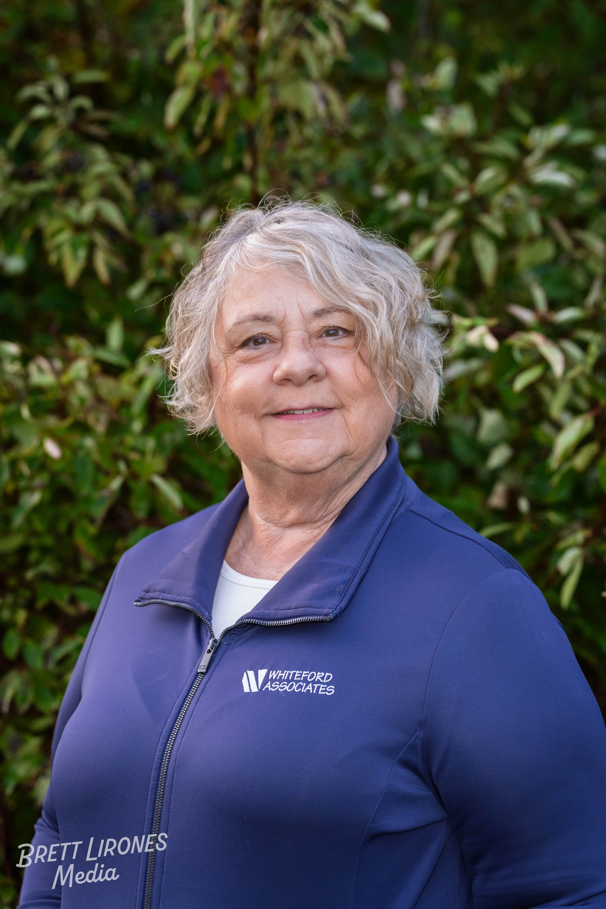 A woman with curly, gray hair standing outside in front of green bushes, wearing a navy blue jacket with 'Whiteford Associates' logo.