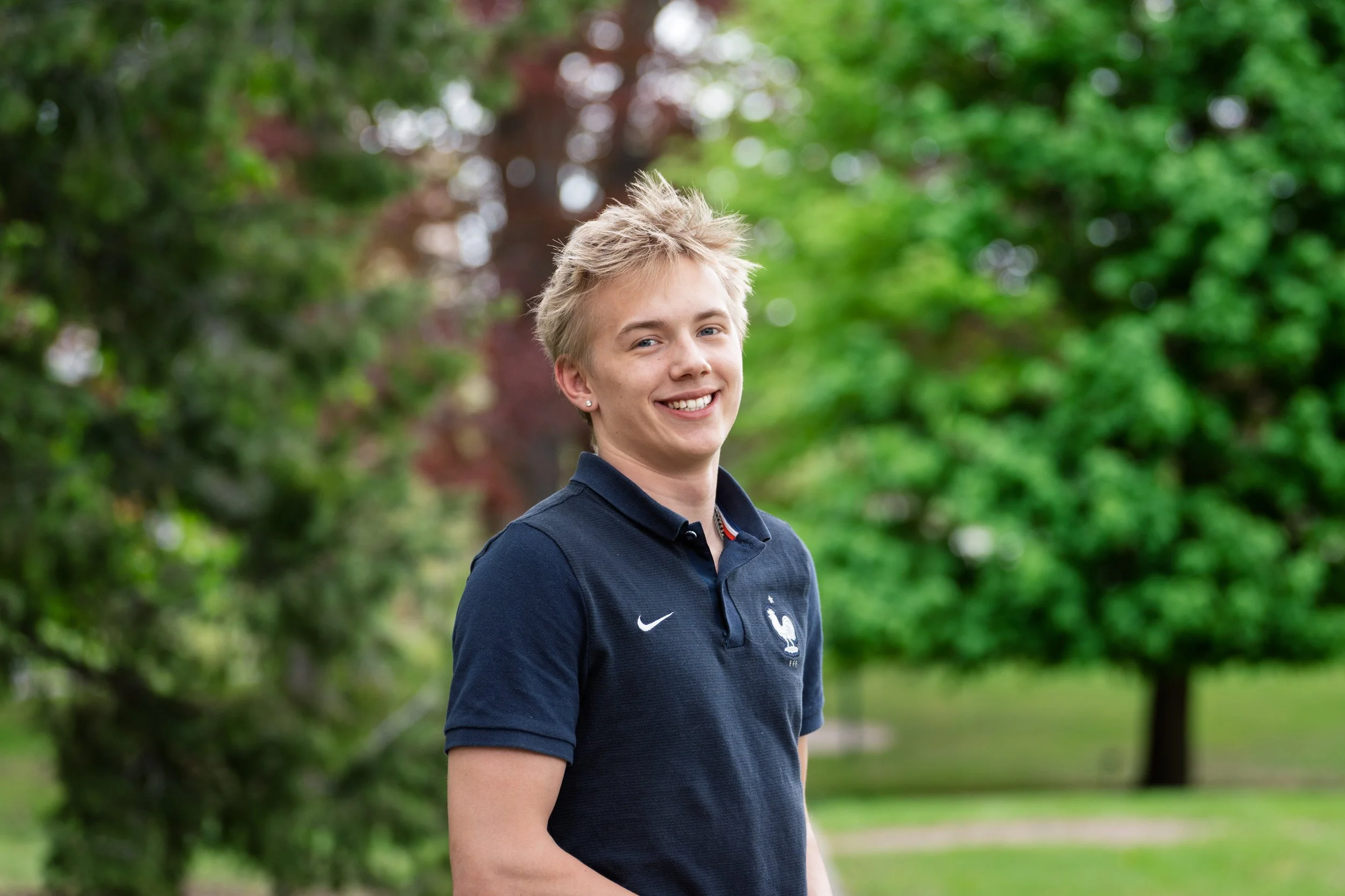 A young man with short, messy blond hair smiling outdoors with green trees in the background, wearing a dark blue sports polo shirt with a French national team emblem.