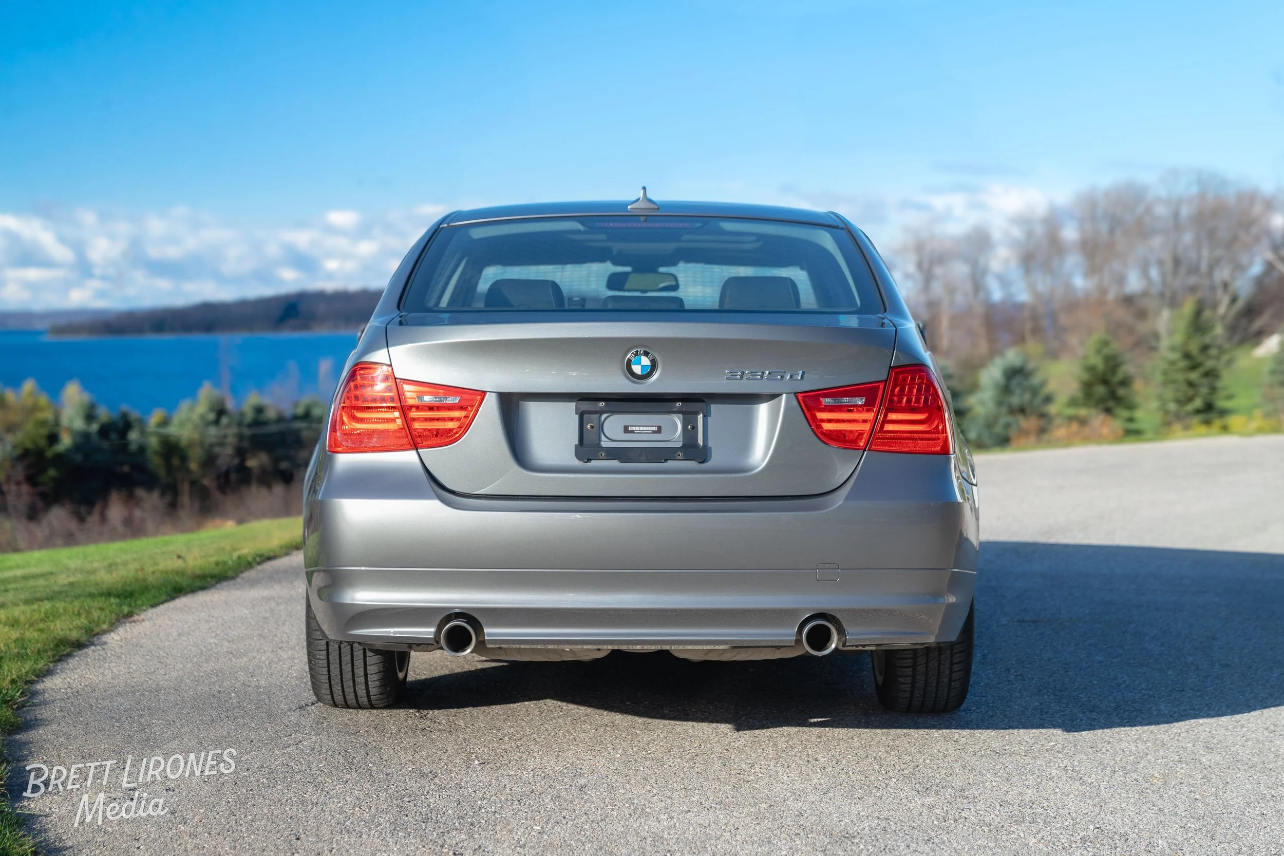 rear view of a silver BMW 3 Series car parked on a road near a grassy area, with a lake, trees, and a blue sky in the background.