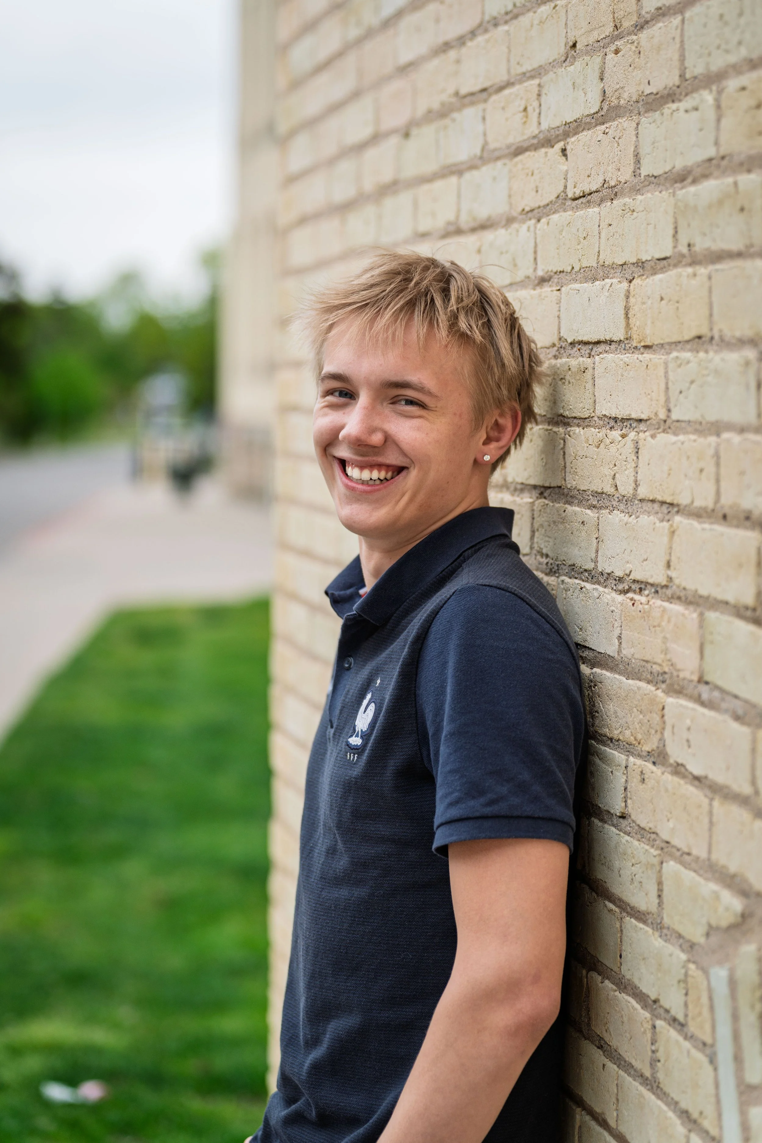 A smiling young man with blond hair, wearing a navy blue polo shirt, leaning against a beige brick wall outdoors.