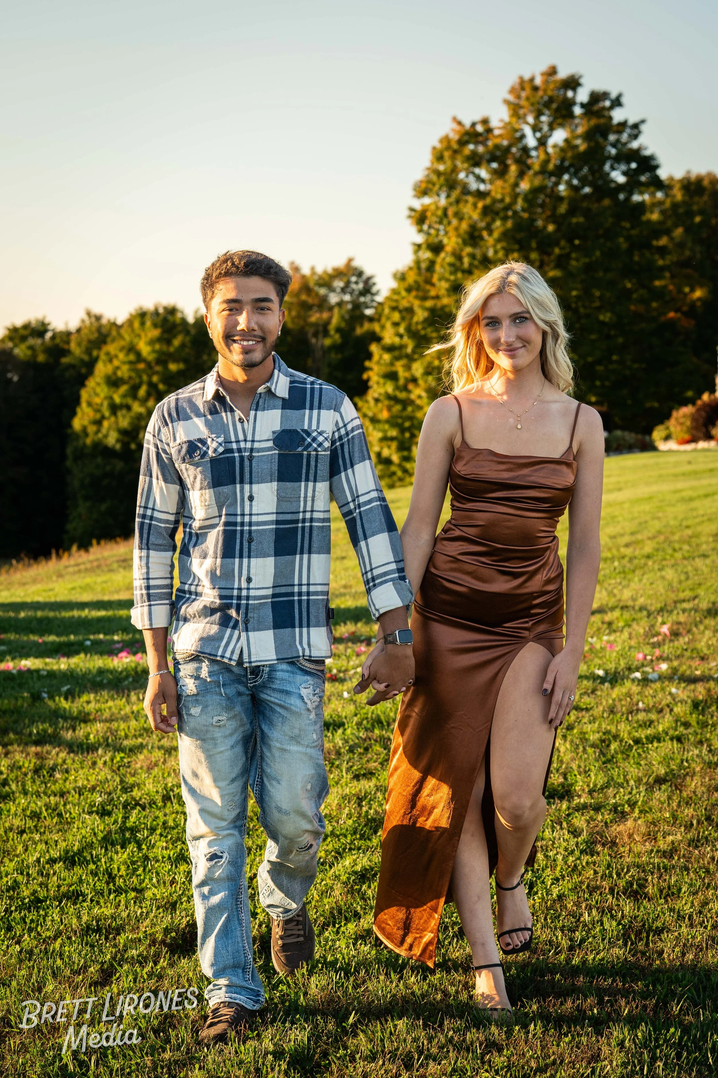 A young couple holding hands walking on a grassy field during sunset. The man wears a plaid shirt and ripped jeans, and the woman wears a brown dress with a thigh-high slit and black heels.