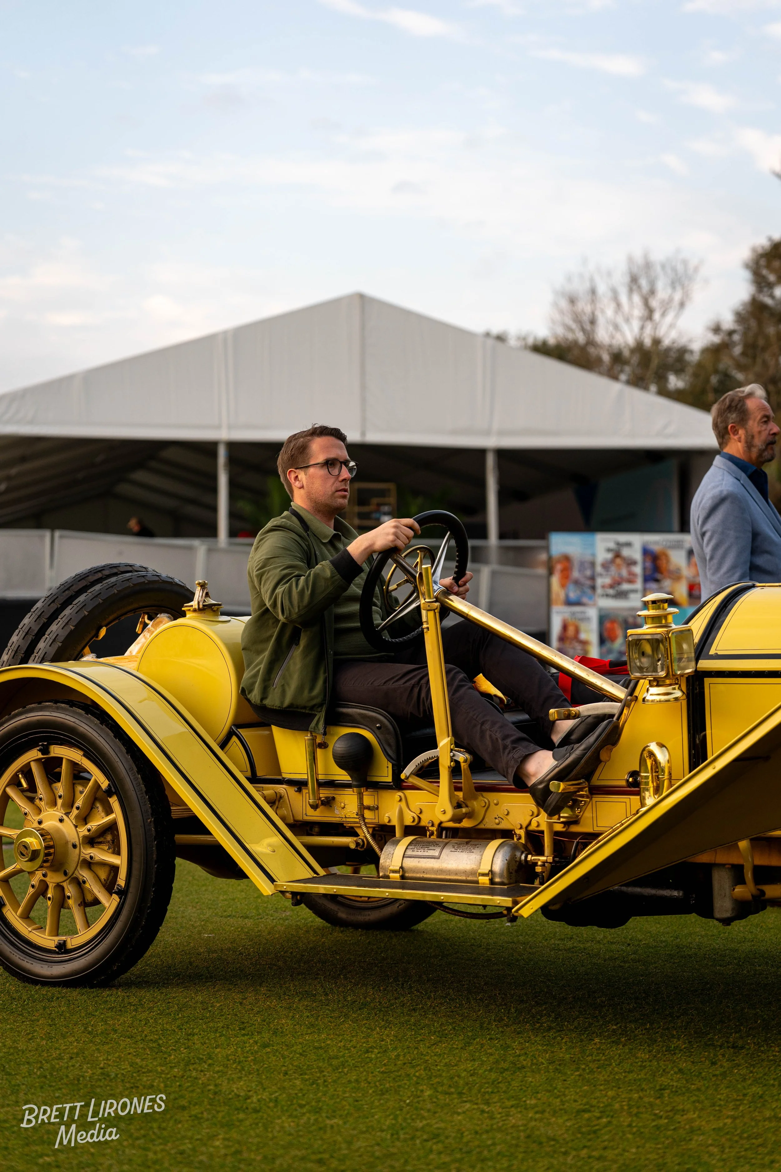 Man sitting in a vintage yellow car during an outdoor event, with a large white tent and some posters visible in the background.