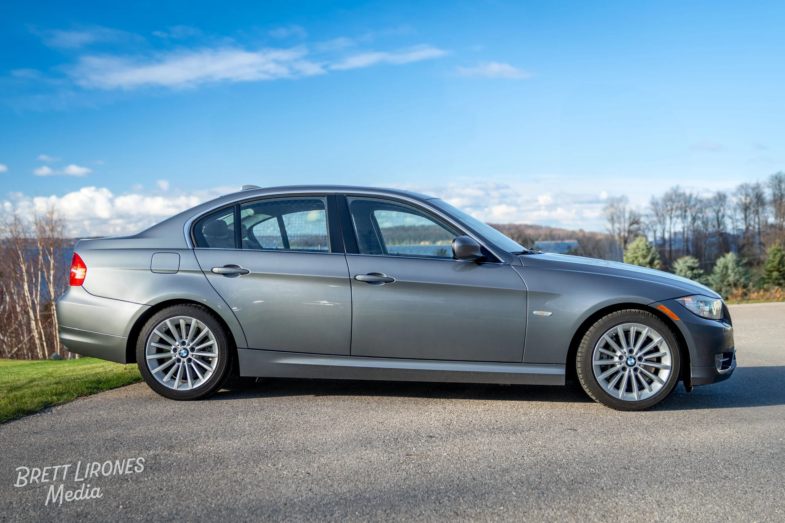 A silver BMW sedan parked on pavement with a scenic background of trees, blue sky, and clouds.