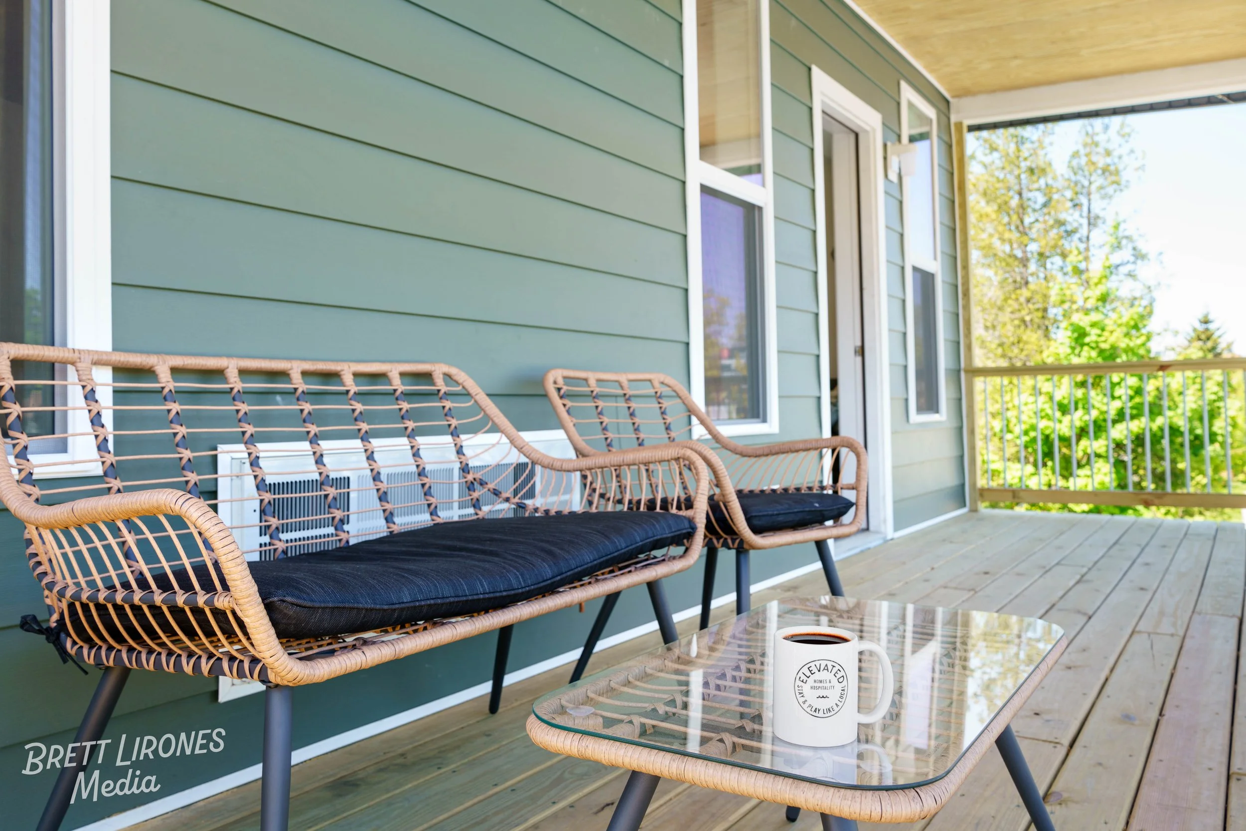 A porch with green siding and a railing, featuring wicker chairs with black cushions, a glass-top table with a white coffee mug, and a small plant. Outside, trees with green leaves are visible.