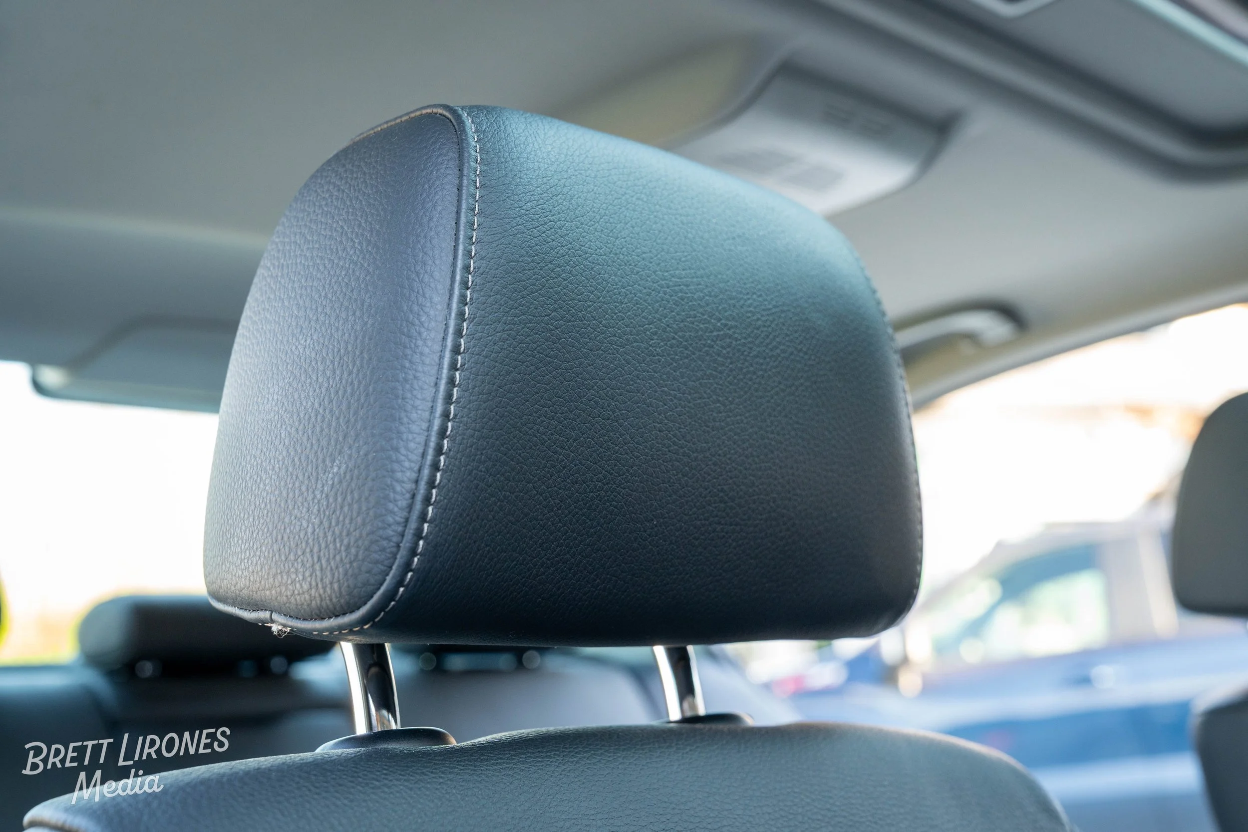 Close-up of a black leather car headrest with stitched seams inside a vehicle, blurred dashboard and car seats in the background.