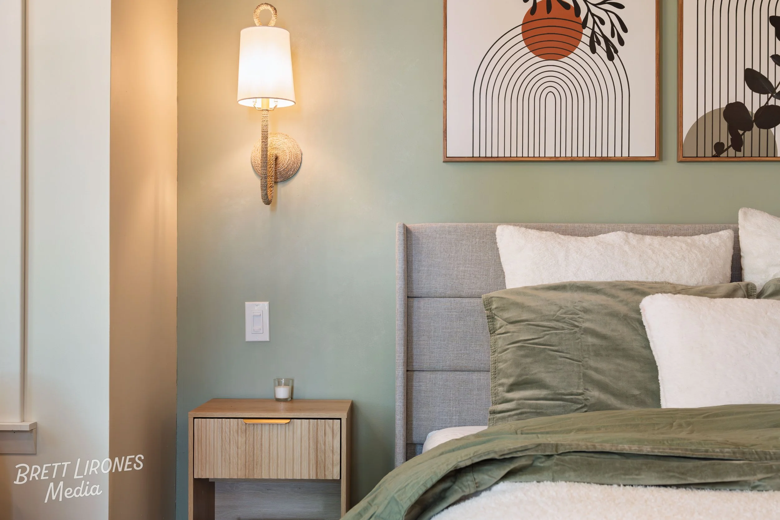 A bedroom corner with a grey upholstered bed, white and green pillows, a small wooden nightstand with a candle, a wall lamp, and two framed abstract art prints on a green wall.