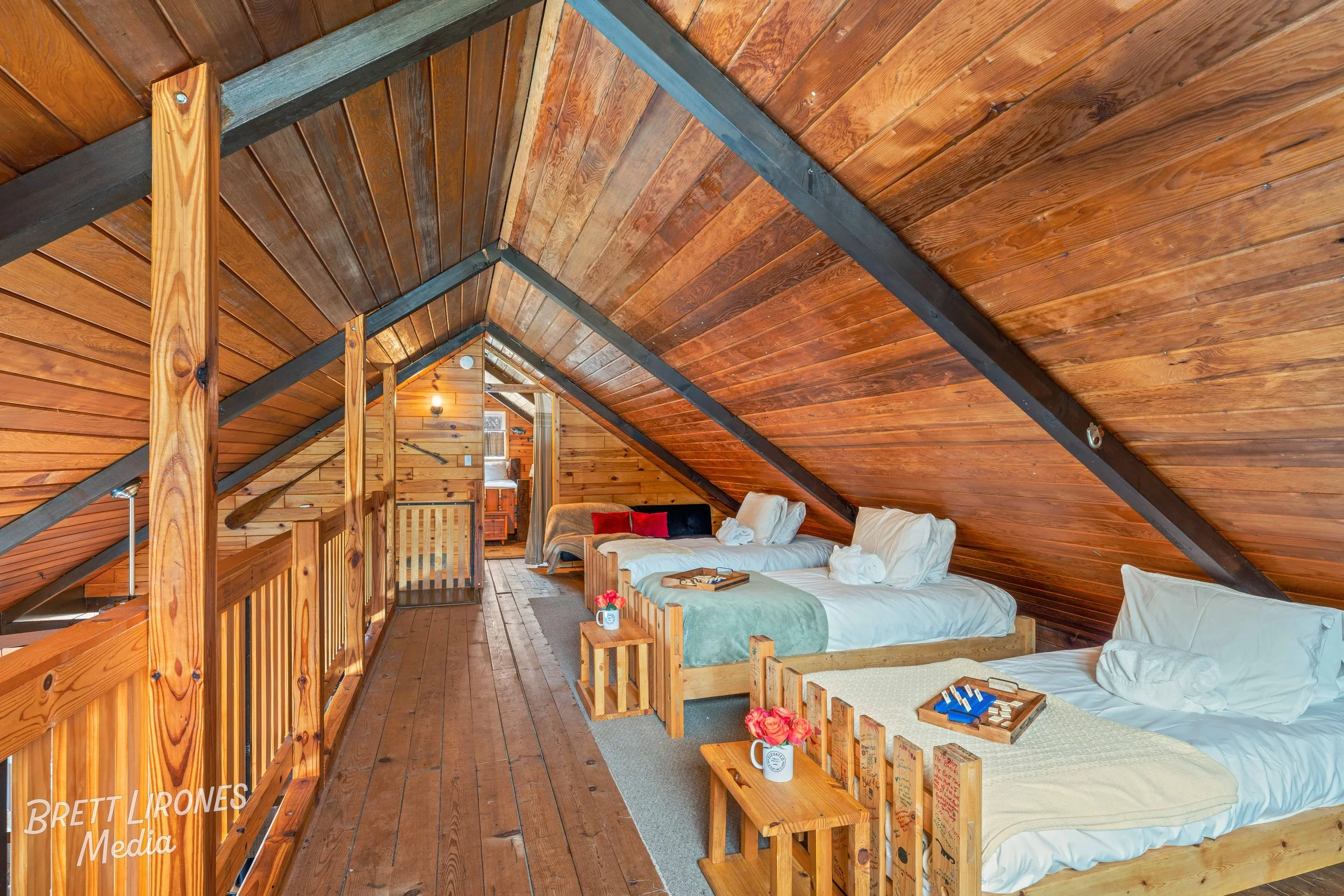Three neatly made beds with white linens in a cozy attic room with sloped wooden ceiling and walls, decorated with small side tables holding vases of pink flowers, and a small sitting area in the background.