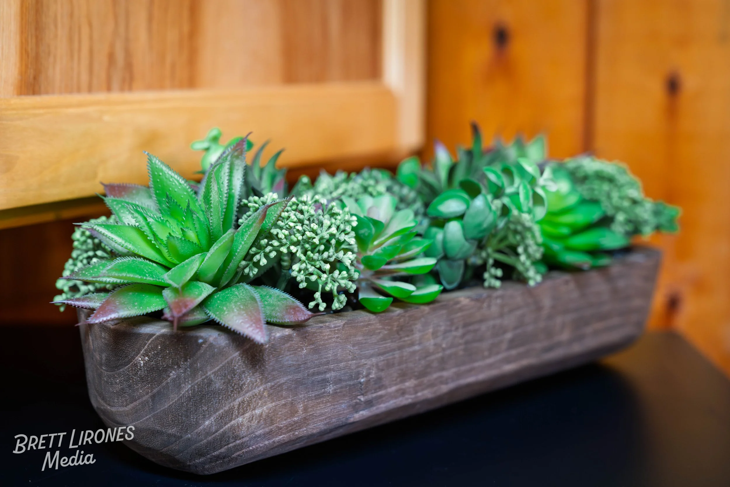 A rectangular wooden planter filled with green and purple succulents, placed on a dark surface with a wood-paneled wall background.