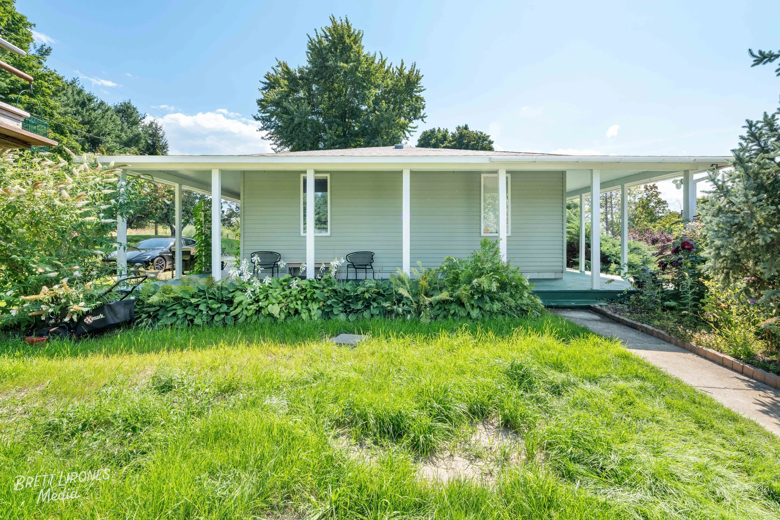 A house with a covered porch, gray siding, and a green lawn in front, surrounded by lush bushes and trees on a sunny day.