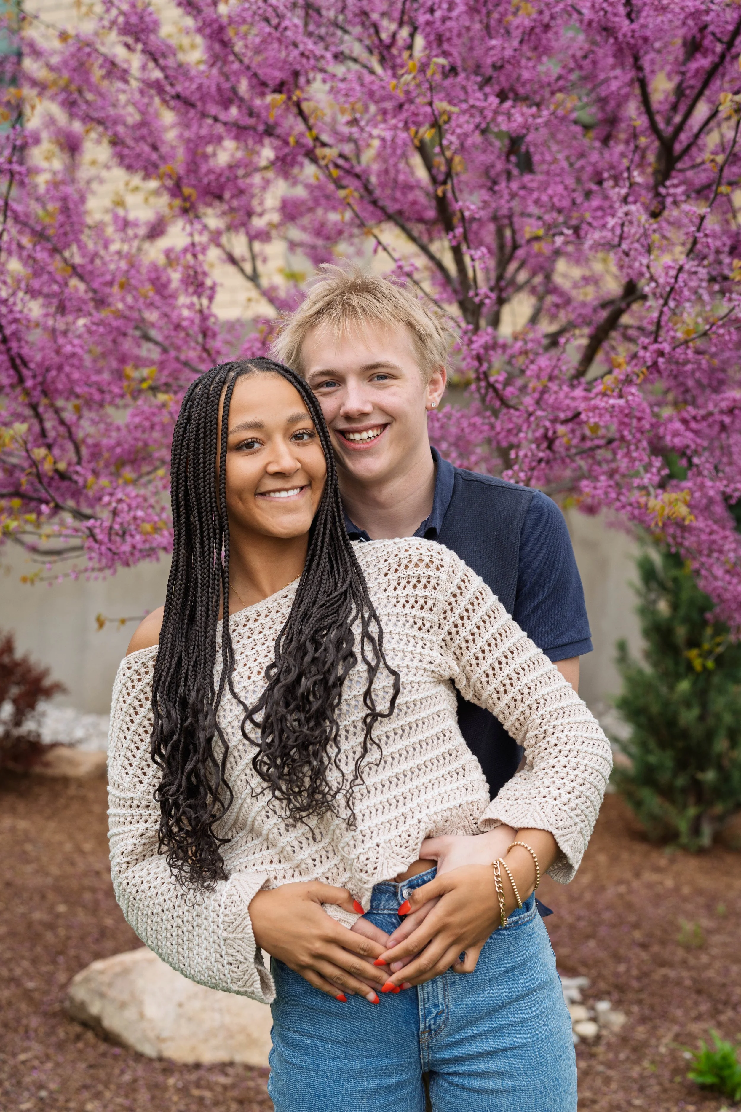 A smiling young woman with long braided hair embracing a young man in front of a blooming pink flowering tree.