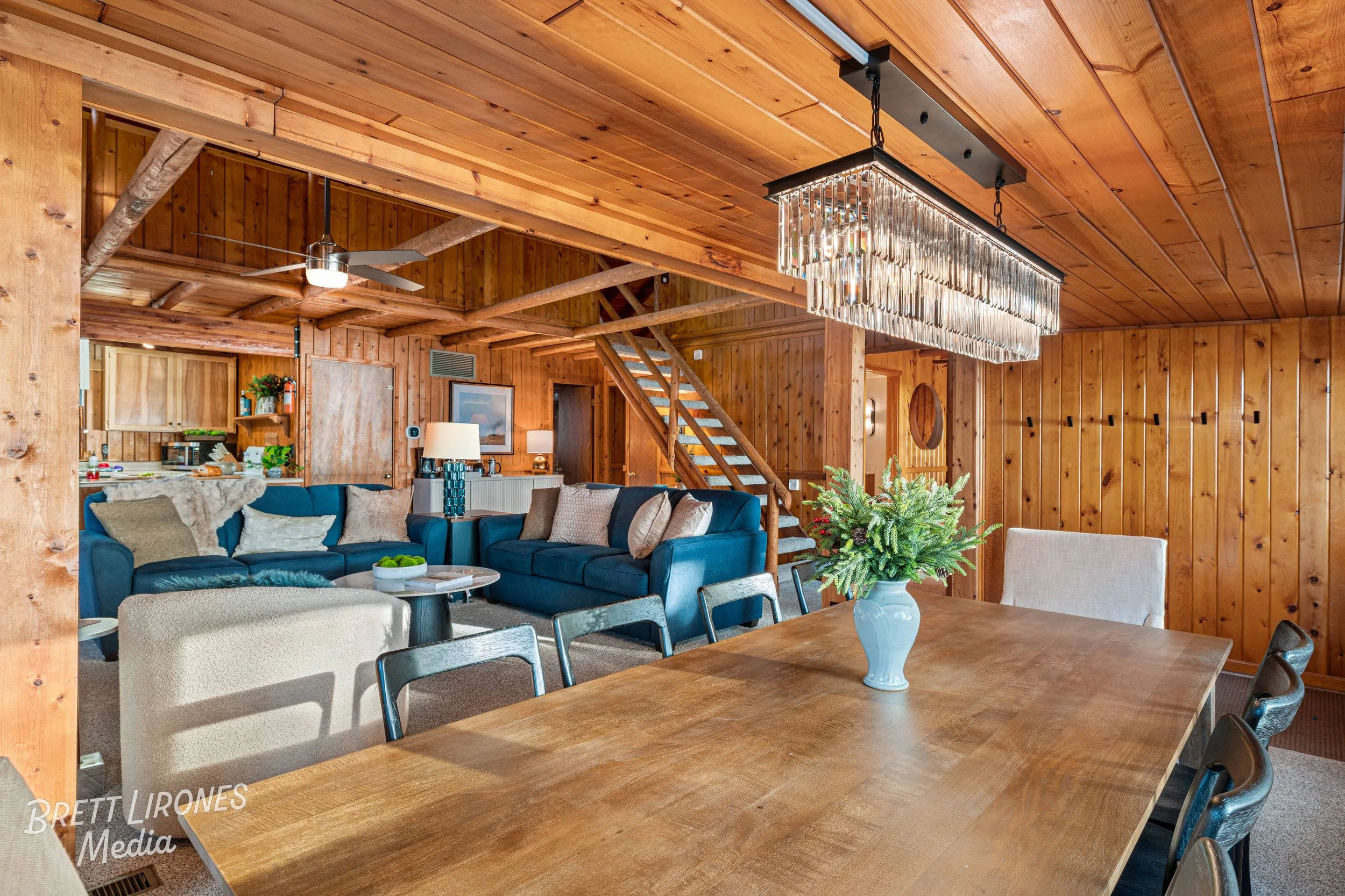 Interior of a cozy, wood-paneled living and dining area with a large wooden table, a white vase with greenery, a blue sofa, and a chandelier hanging from the ceiling.