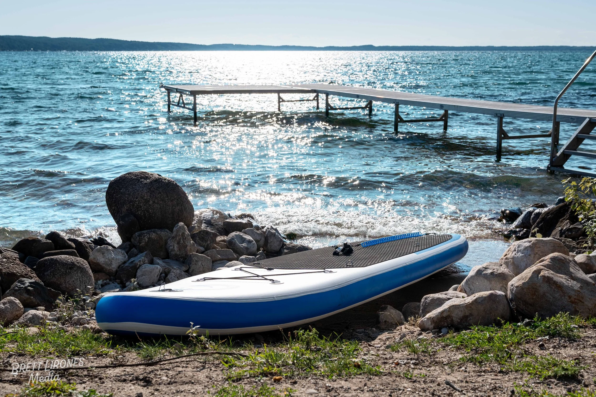 A paddleboard resting on rocks on a lakeshore with a dock extending into the water and shimmering sunlight on the lake.