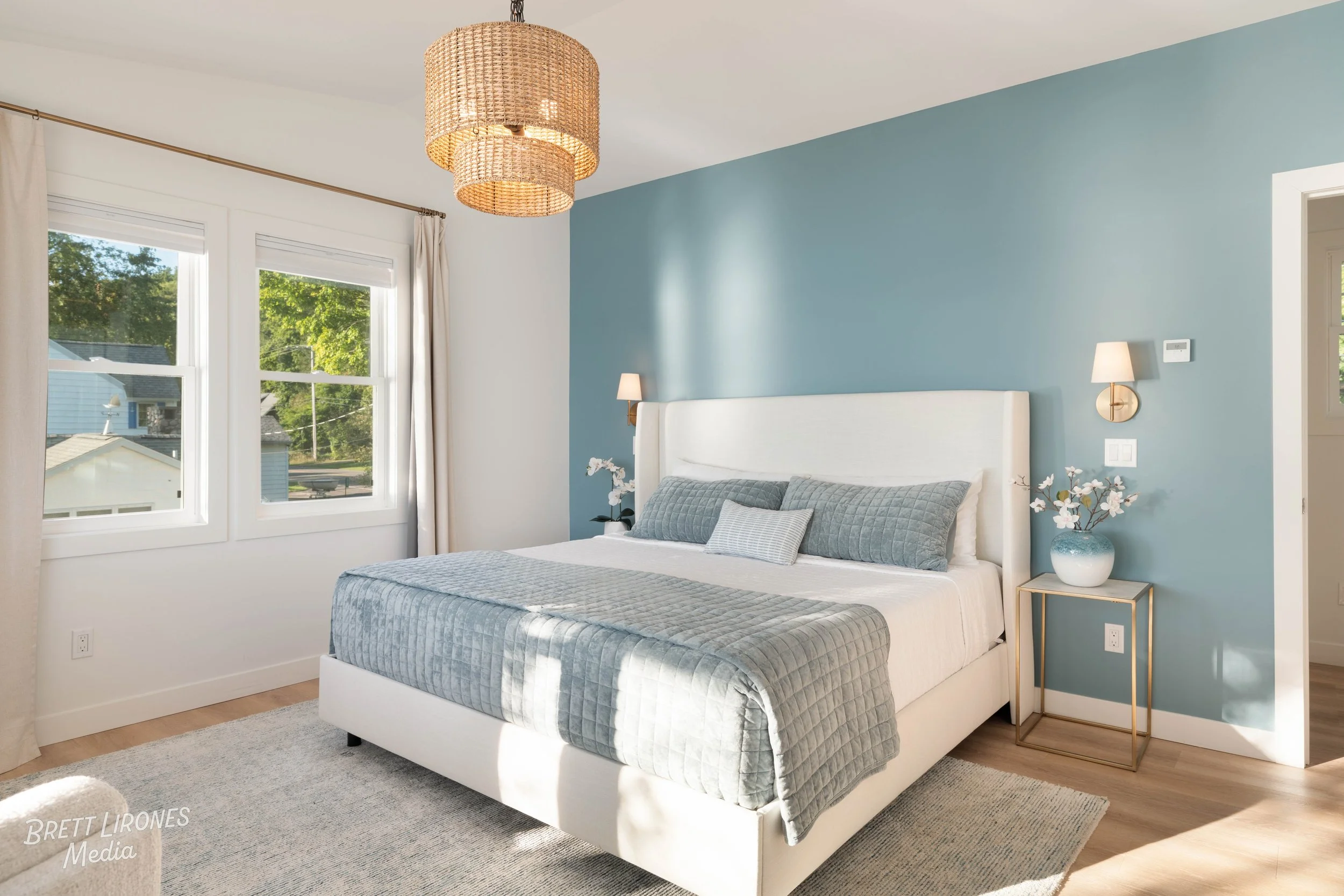 A bedroom with a white bed, blue accent wall, two wall-mounted lamps, nightstand with a flower vase, and natural light from windows.