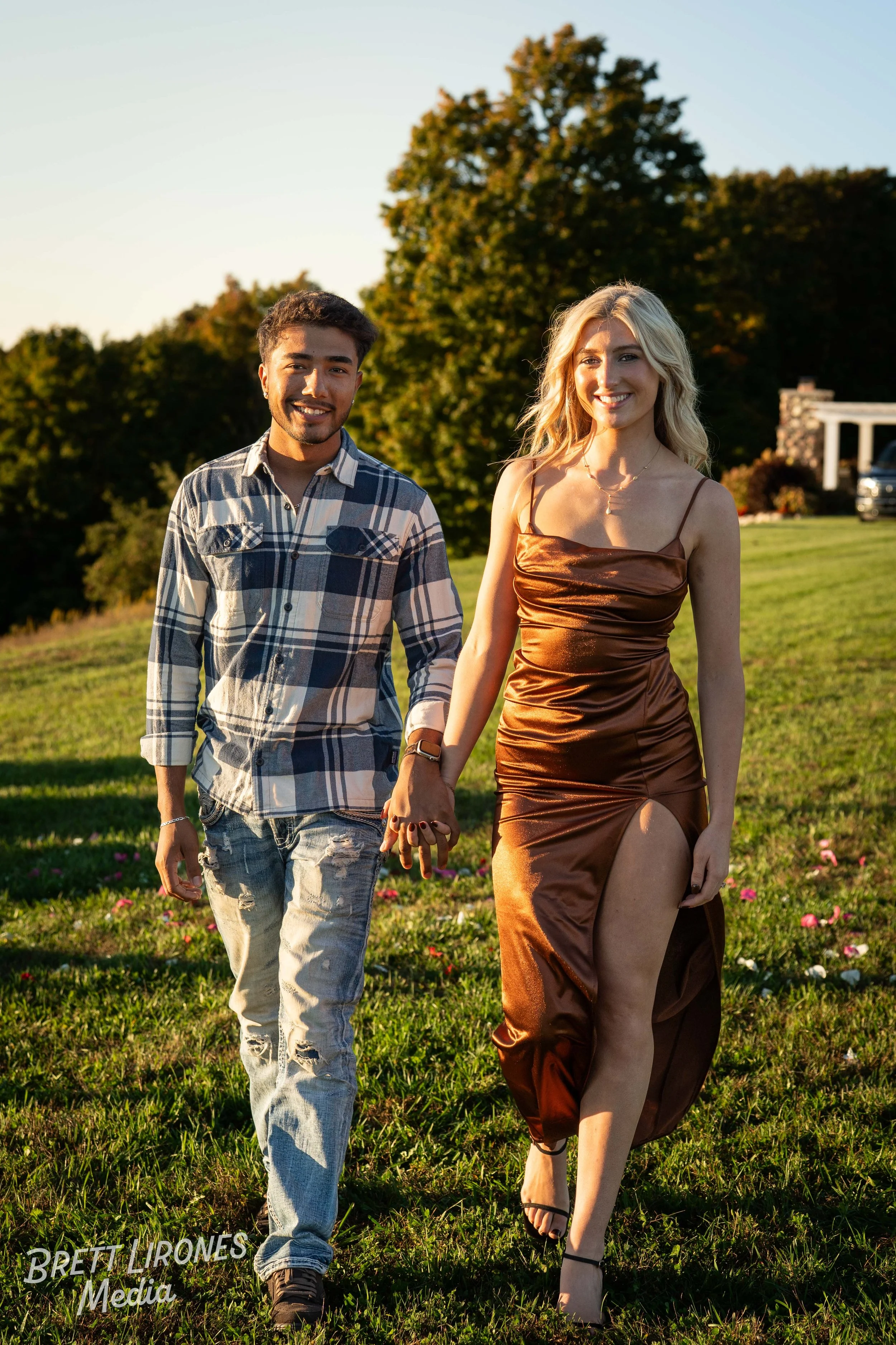 A young man and woman walking hand in hand outdoors on grassy terrain with trees and a house in the background, smiling at the camera during sunset.