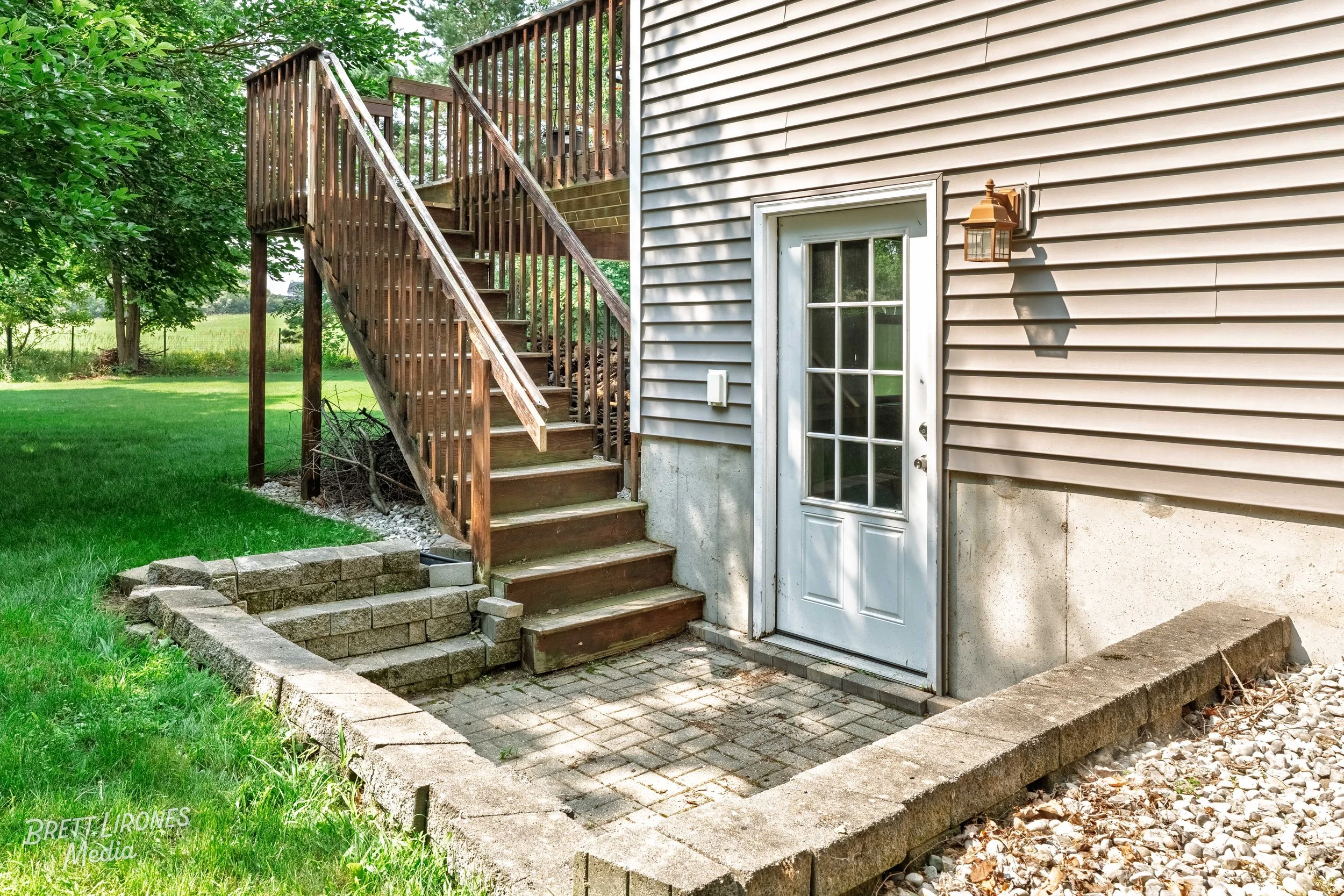 Backyard with wooden staircase leading to a deck, concrete and brick patio area with a white door, exterior siding of house, outdoor light fixture, green grass, and trees in background.