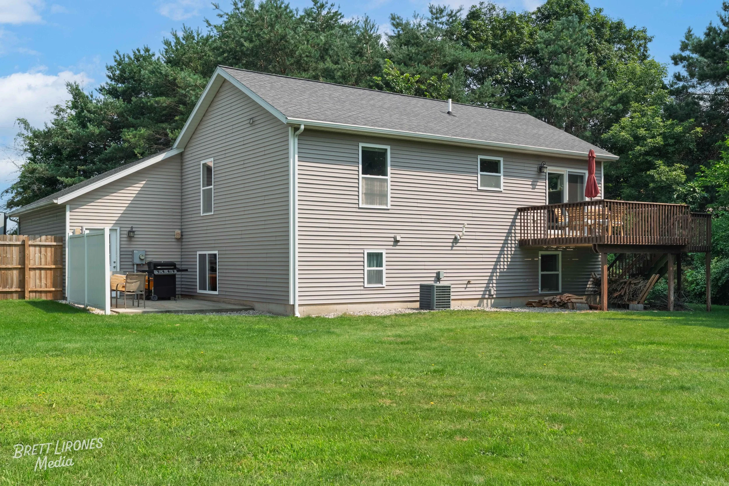 Backyard view of a two-story house with beige siding, a wooden deck with patio furniture and an umbrella, surrounded by green grass and trees.