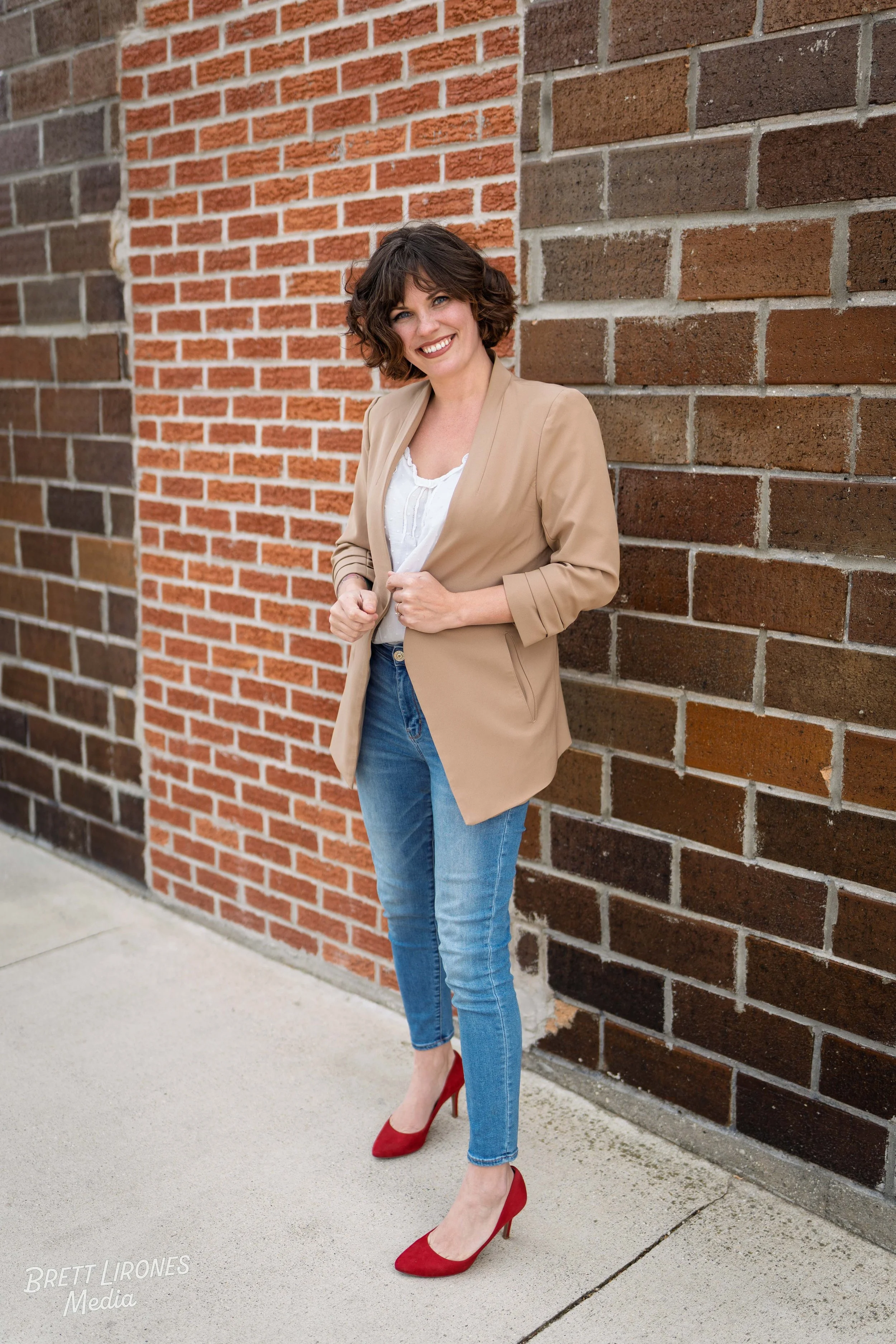 A woman with curly brown hair, wearing a beige blazer, white top, blue jeans, and red high-heeled shoes, standing outdoors against a brick wall, smiling at the camera.