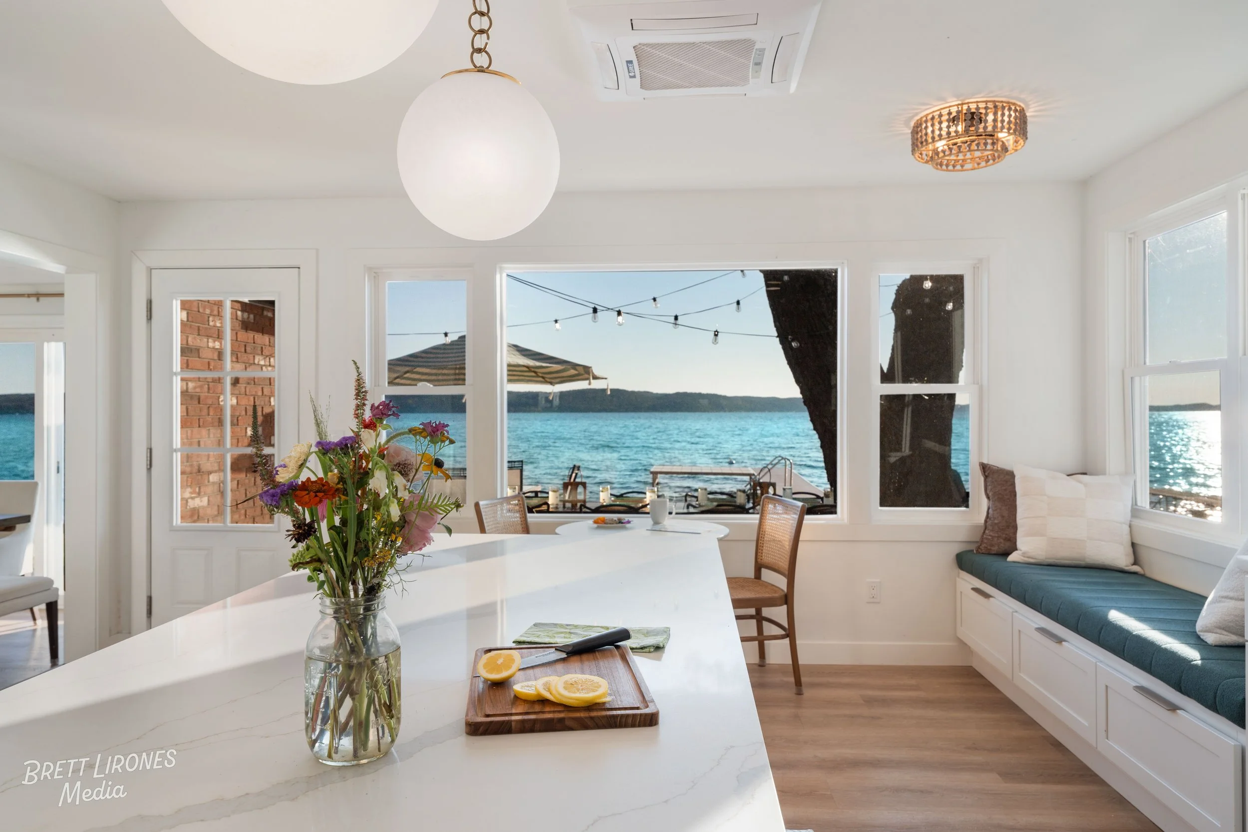 Interior view of a bright dining area with large windows overlooking a lake, featuring a white counter with a vase of flowers and lemon slices, string lights outside, and a view of boat docks and umbrellas.