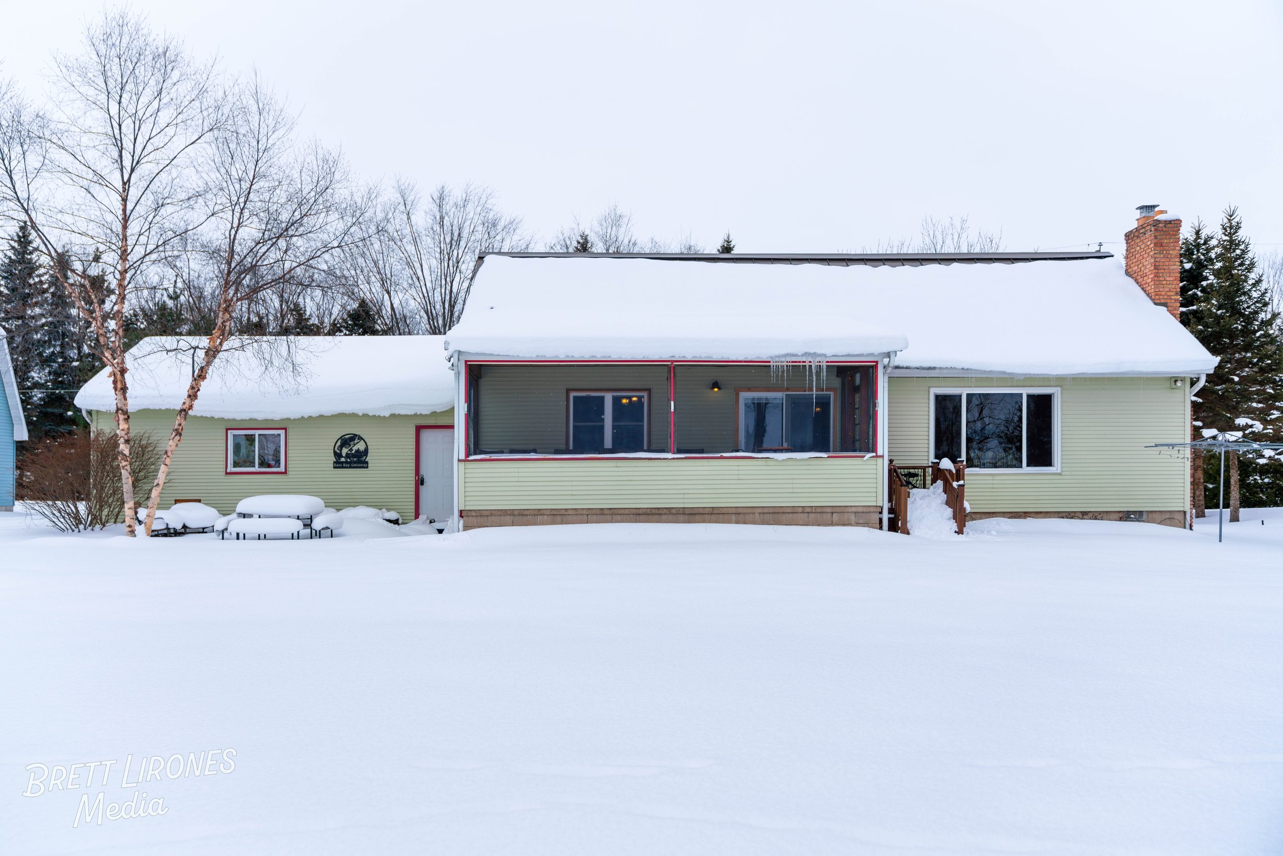 A yellow house with a snow-covered roof and a screened porch in a snowy landscape with trees in the background.