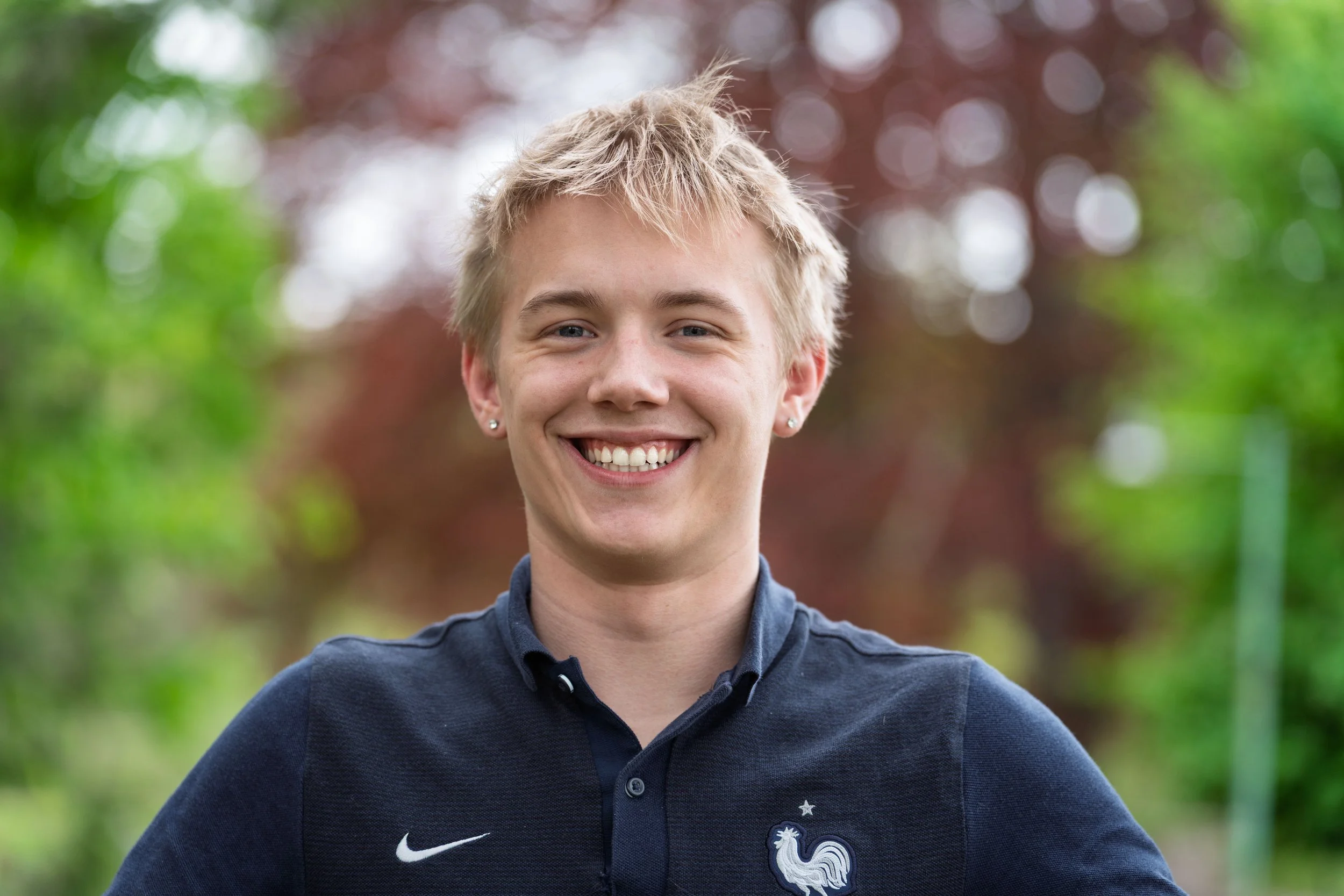 Smiling young man with short blonde hair wearing a navy blue shirt with a French football team logo, standing outdoors with trees and blurred background.