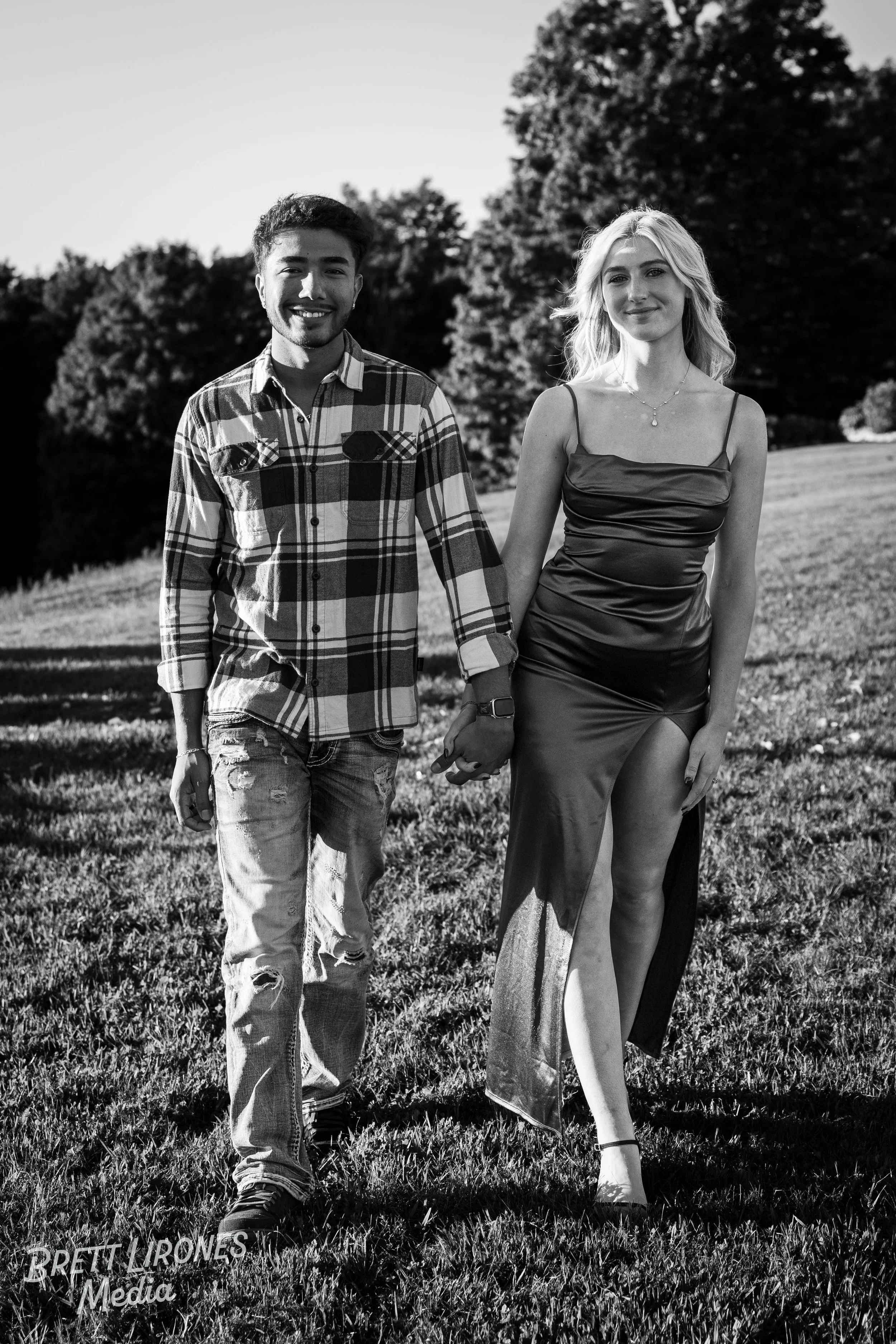 A black and white photo of a smiling young man holding hands with a young woman in a fancy dress, walking outdoors in a park with trees in the background.