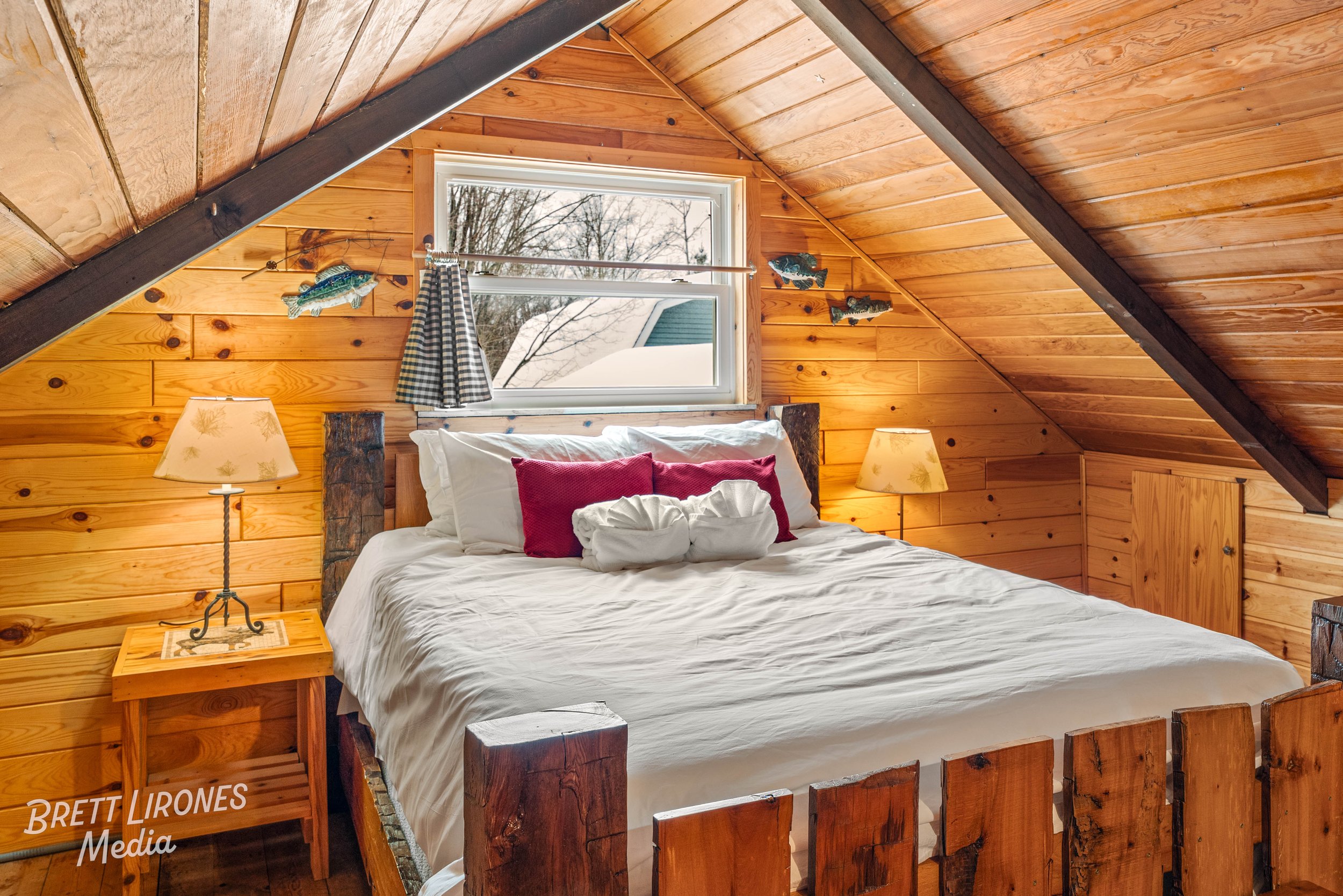 A cozy bedroom with wooden paneled walls and sloped ceiling, featuring a bed with white linens, red pillows, and two swan-shaped towel arrangements, flanked by two bedside lamps, and decorated with mounted fish and a window showing an outdoor winter 