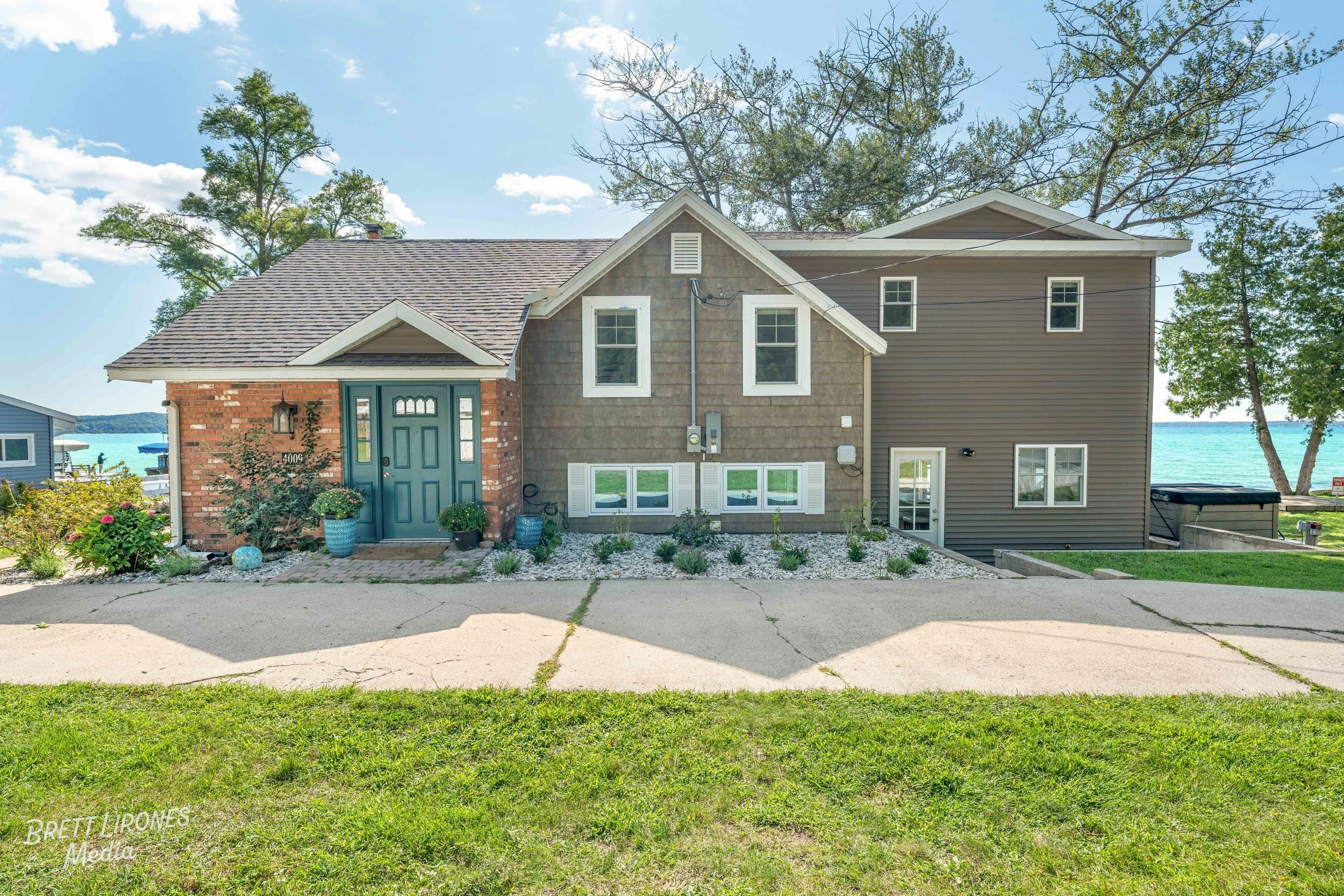 A two-story house with a brick and gray siding exterior, blue front door, and windows overlooking a lake in the background. The house has a landscaped garden with potted plants and rocks, and a concrete driveway.