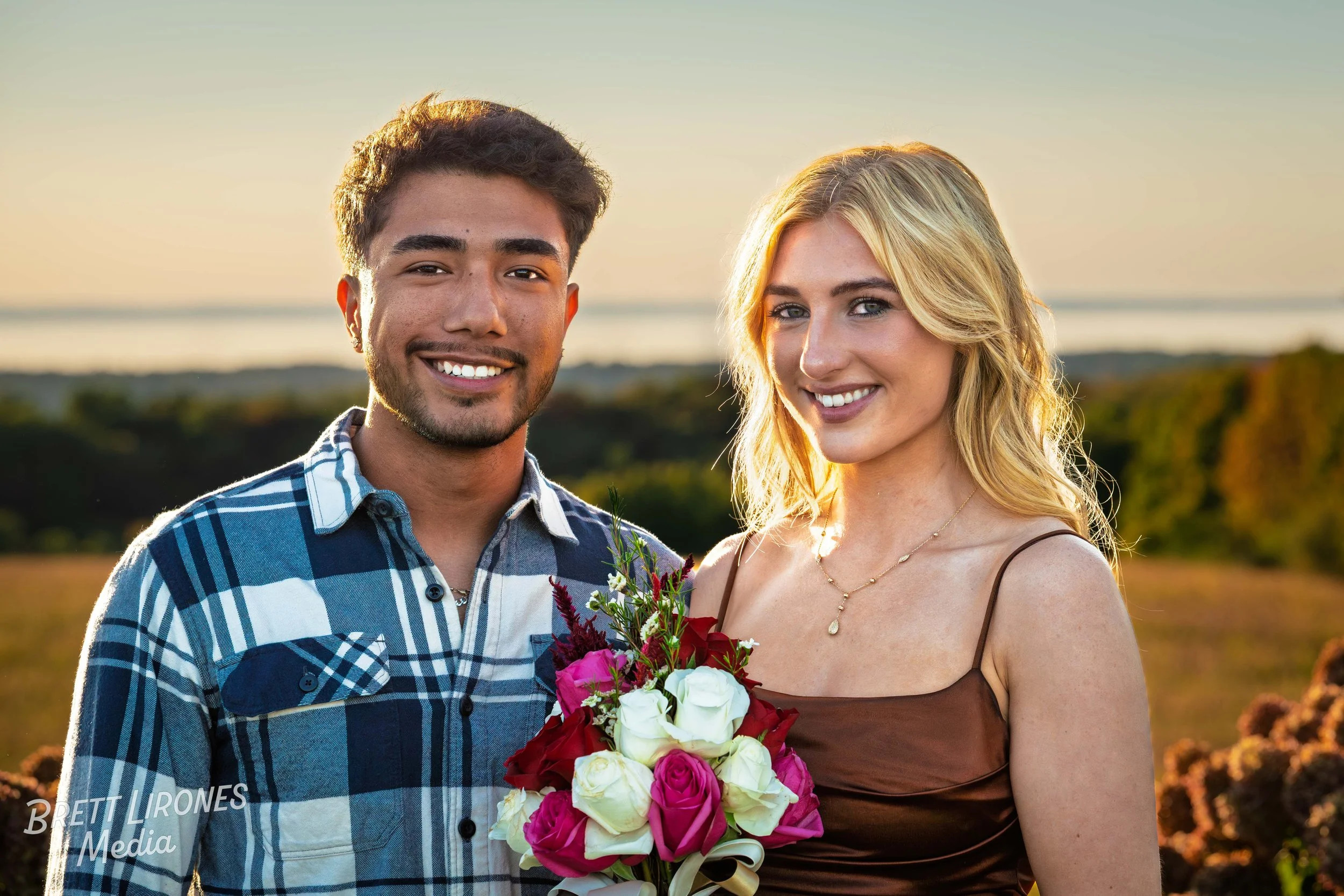 A young man and woman smiling outdoors during sunset, with the woman holding a bouquet of pink, white, and red roses.