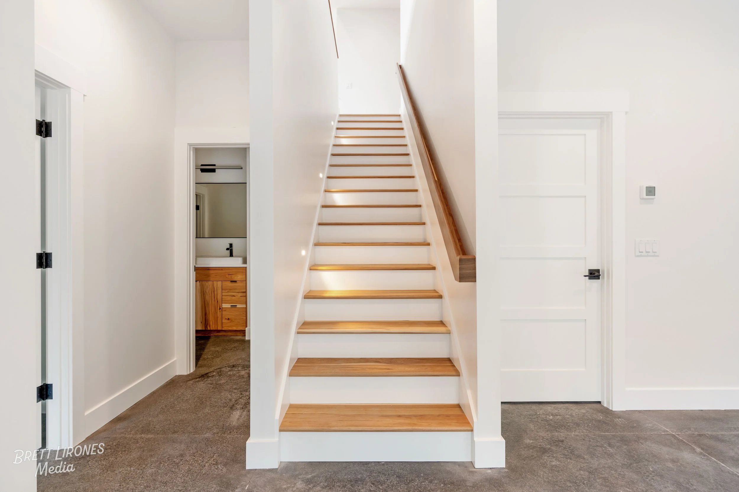 Interior view of a modern home staircase with wooden steps and a wooden handrail, leading upwards with interior design elements like white walls and doors visible.