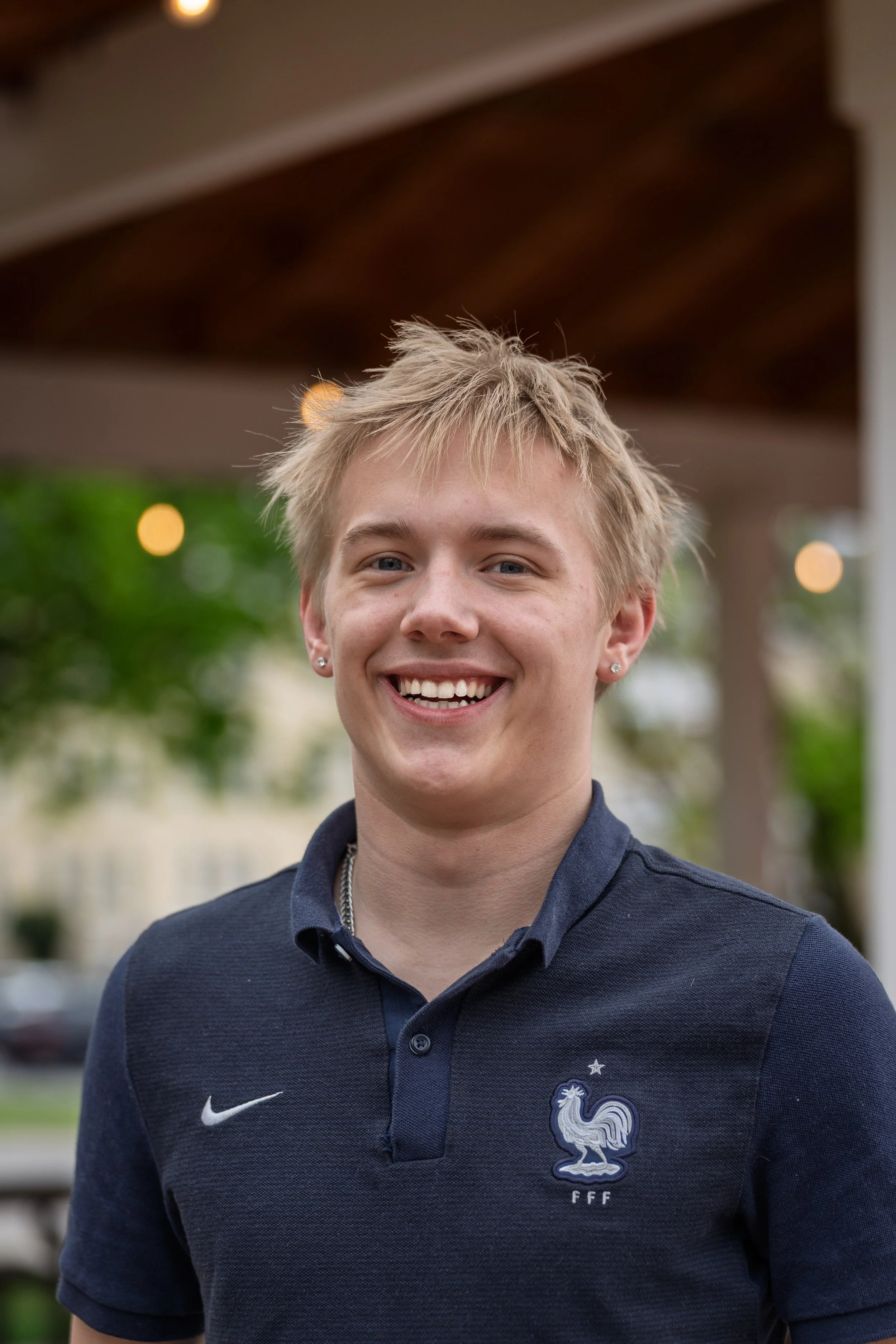 A young man smiling outdoors, wearing a navy blue polo shirt with a French football crest on the chest.