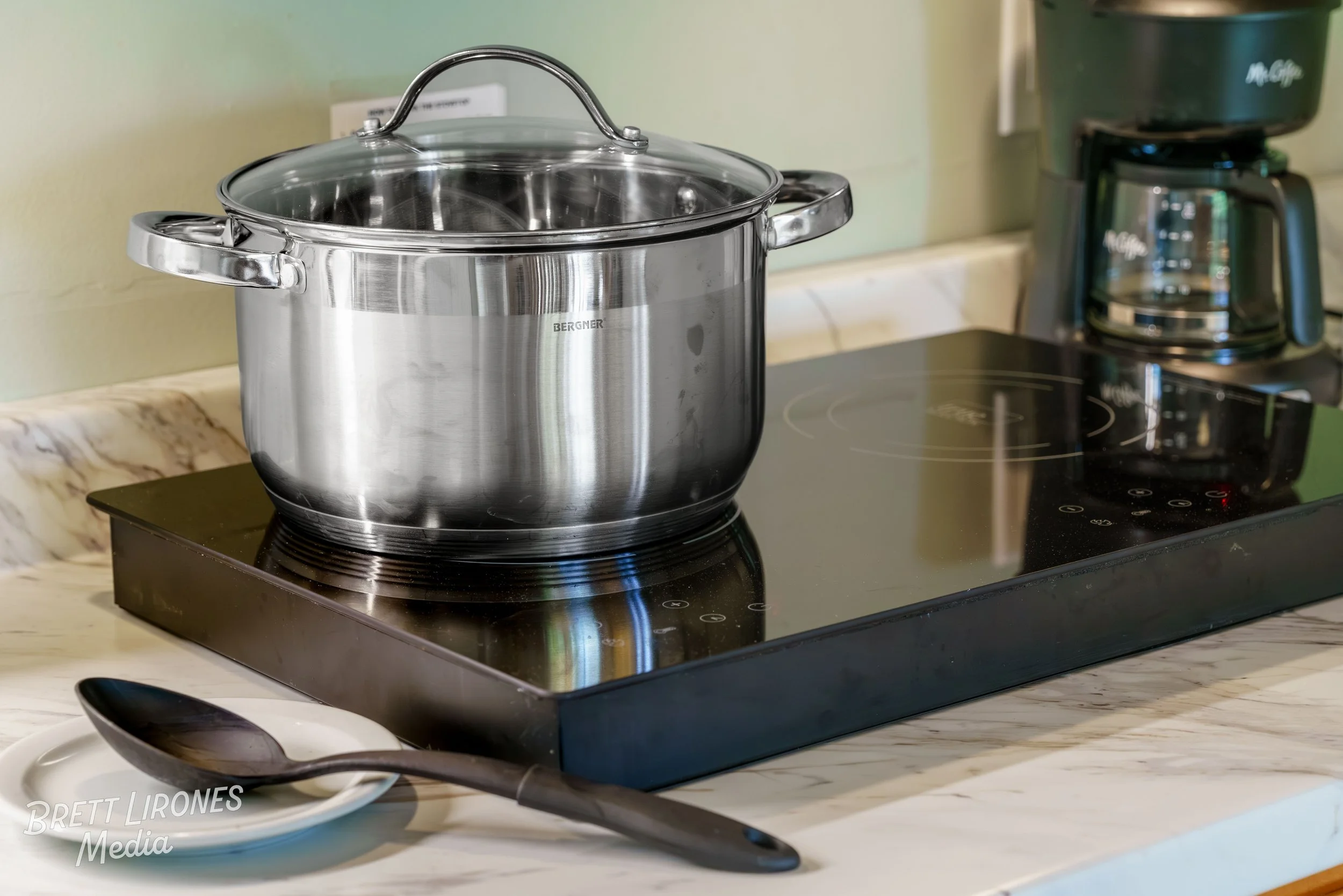A stainless steel pot with a glass lid sits on an electric stove. A black spoon rests on a white plate in front of the stove, and a coffee maker is seen in the background.