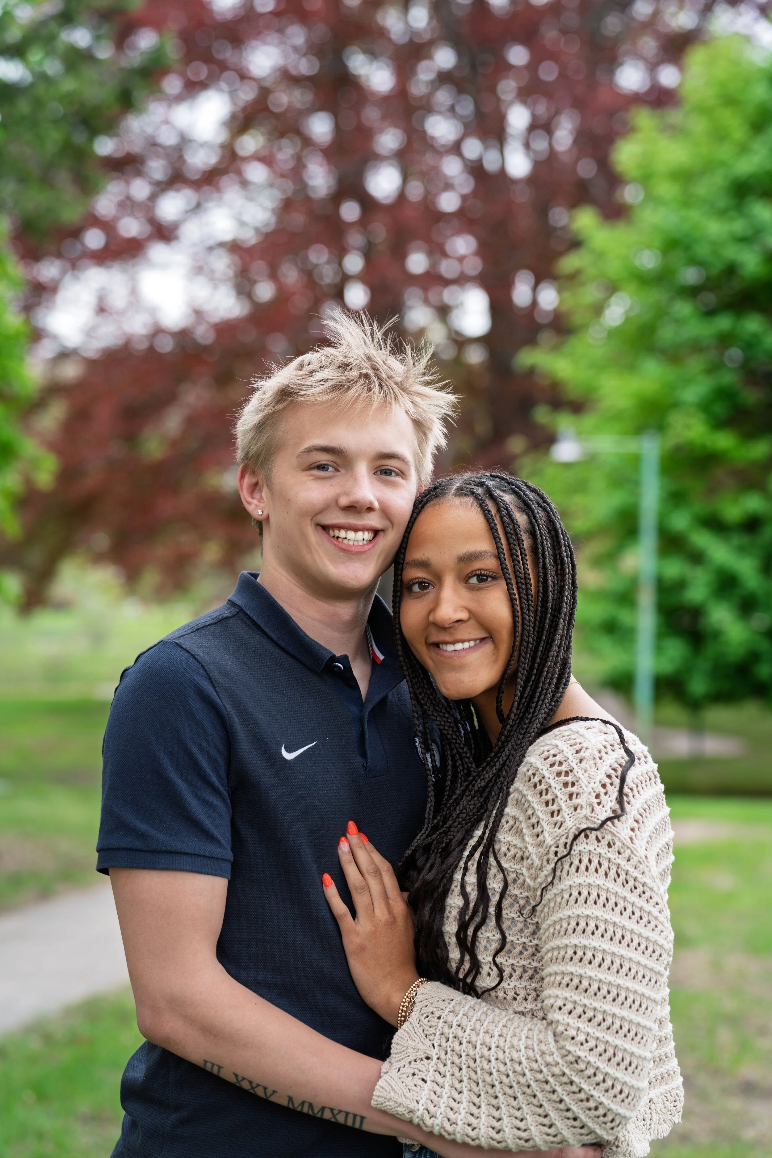 A happy couple smiling outdoors in a park with green and red trees.