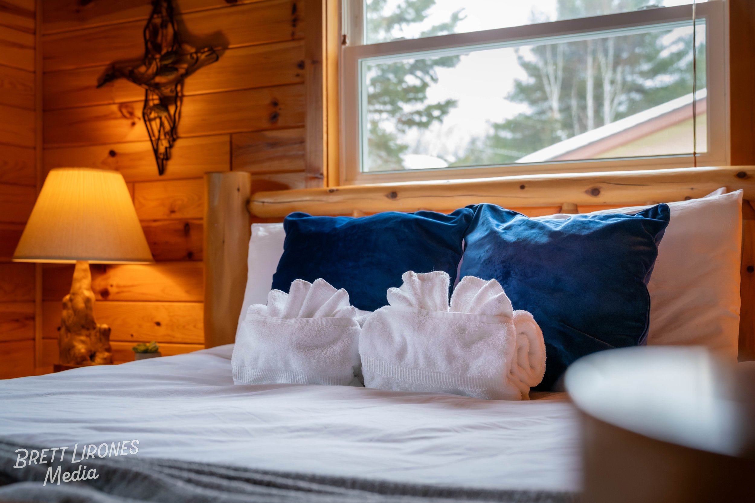 A cozy bedroom with a wooden wall and a window showing an outdoor scene. The bed has a white sheet, two large navy blue pillows, and two neatly rolled white towels placed on it. A warm glowing lamp is on a wooden nightstand beside the bed.
