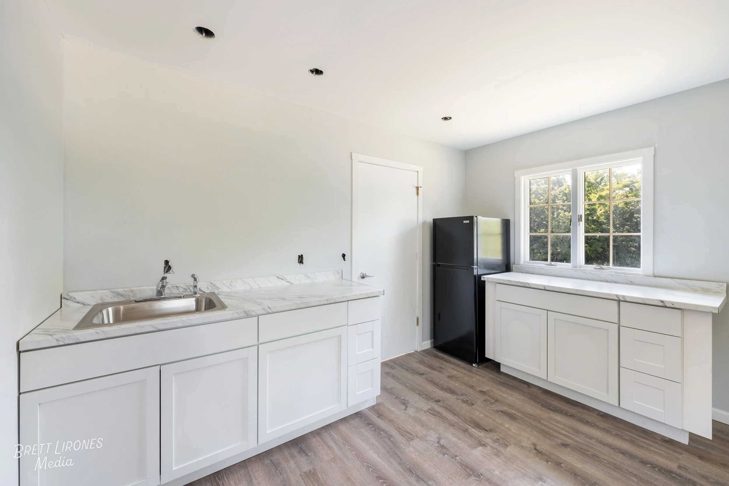 Empty kitchen with white cabinets, marbled countertops, black refrigerator, and a window with trees outside