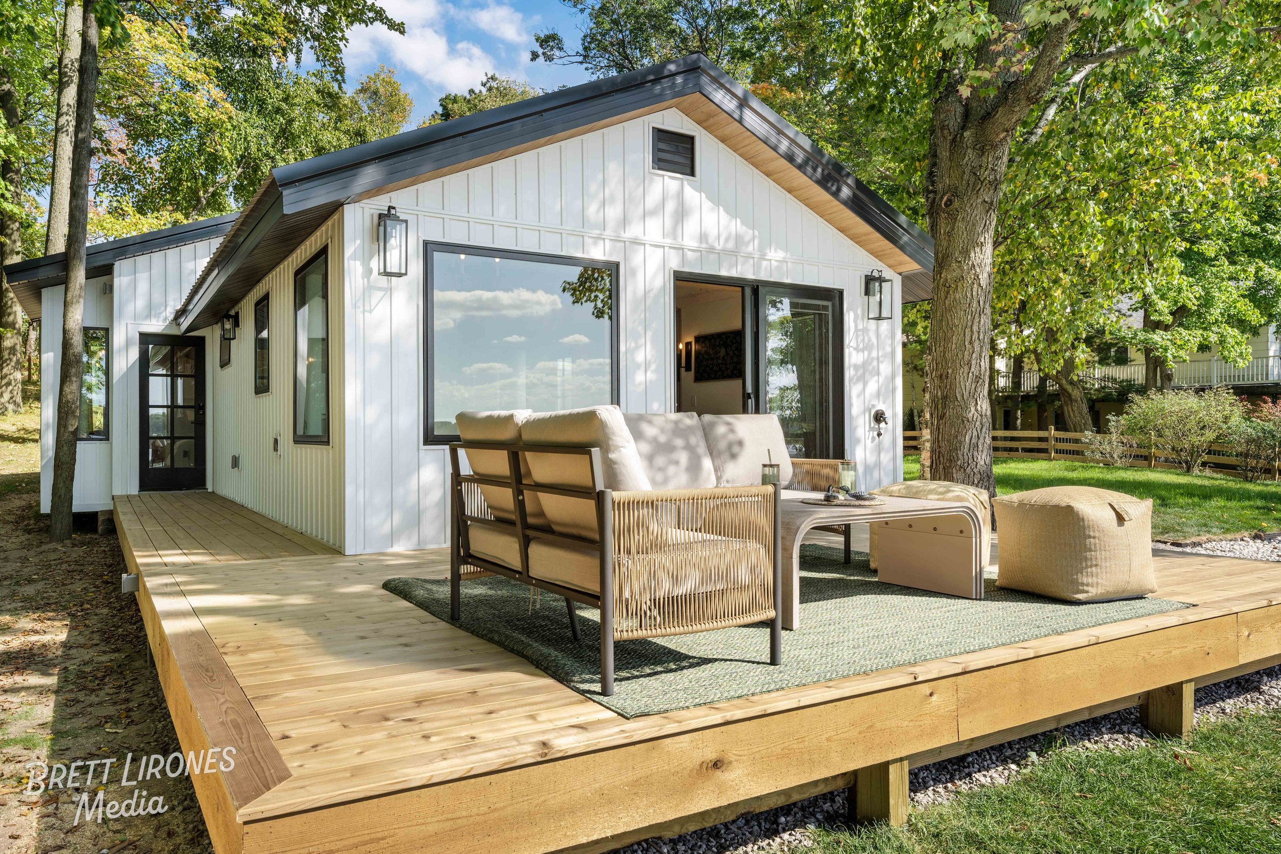A modern white house with large windows and sliding glass doors, surrounded by green trees and a backyard patio with outdoor furniture.