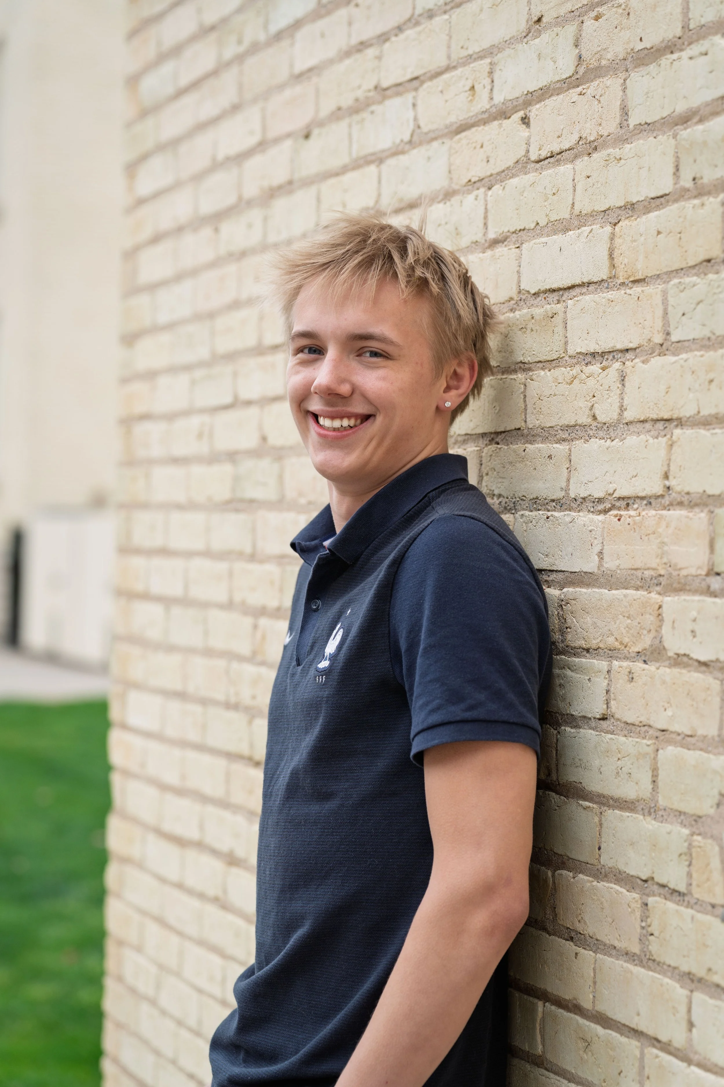Smiling young man leaning against a brick wall outside, wearing a navy polo shirt.