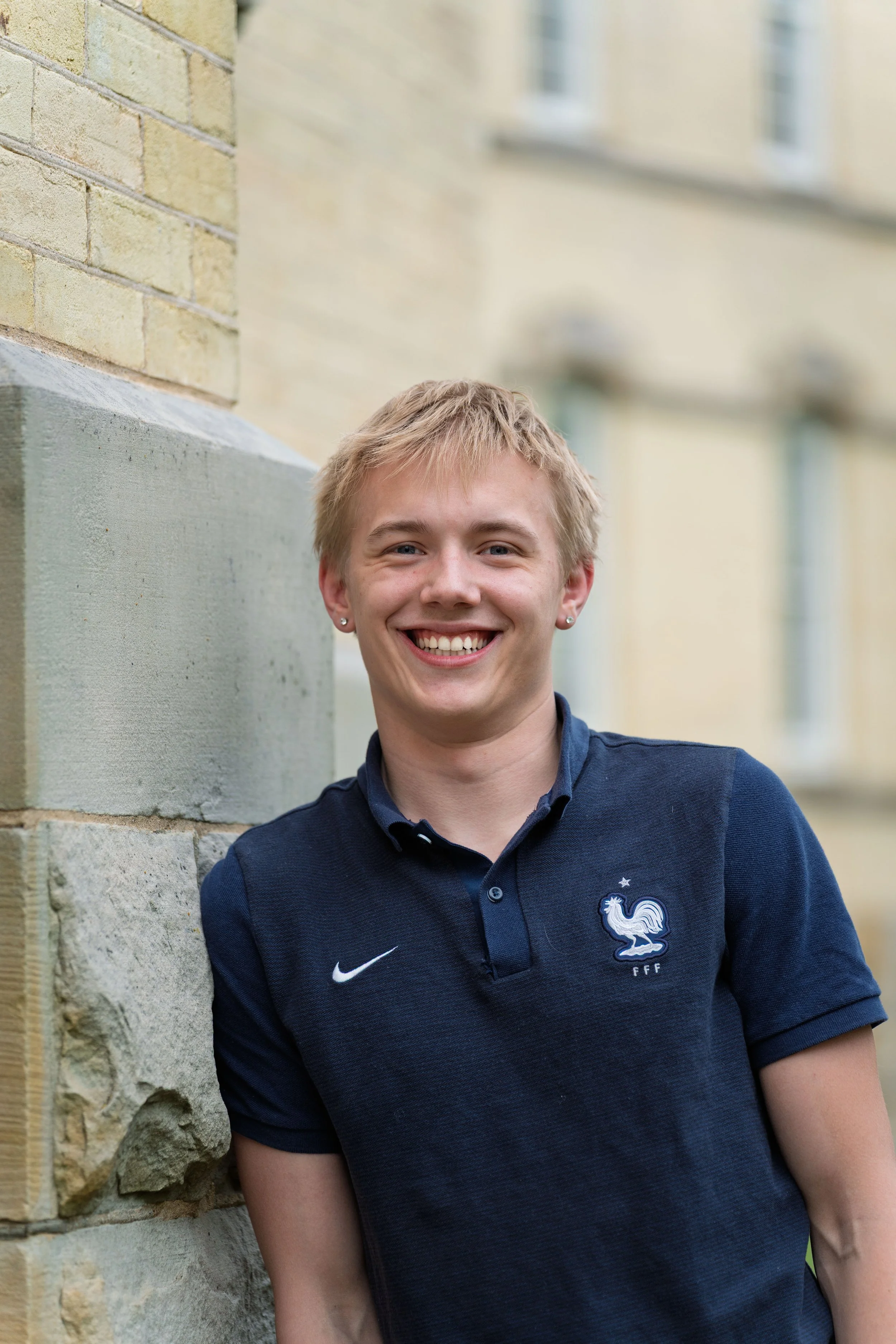 A smiling young man with blond hair and earrings, wearing a navy blue polo shirt with a French football emblem, leaning against a stone wall outdoors with a blurred building background.