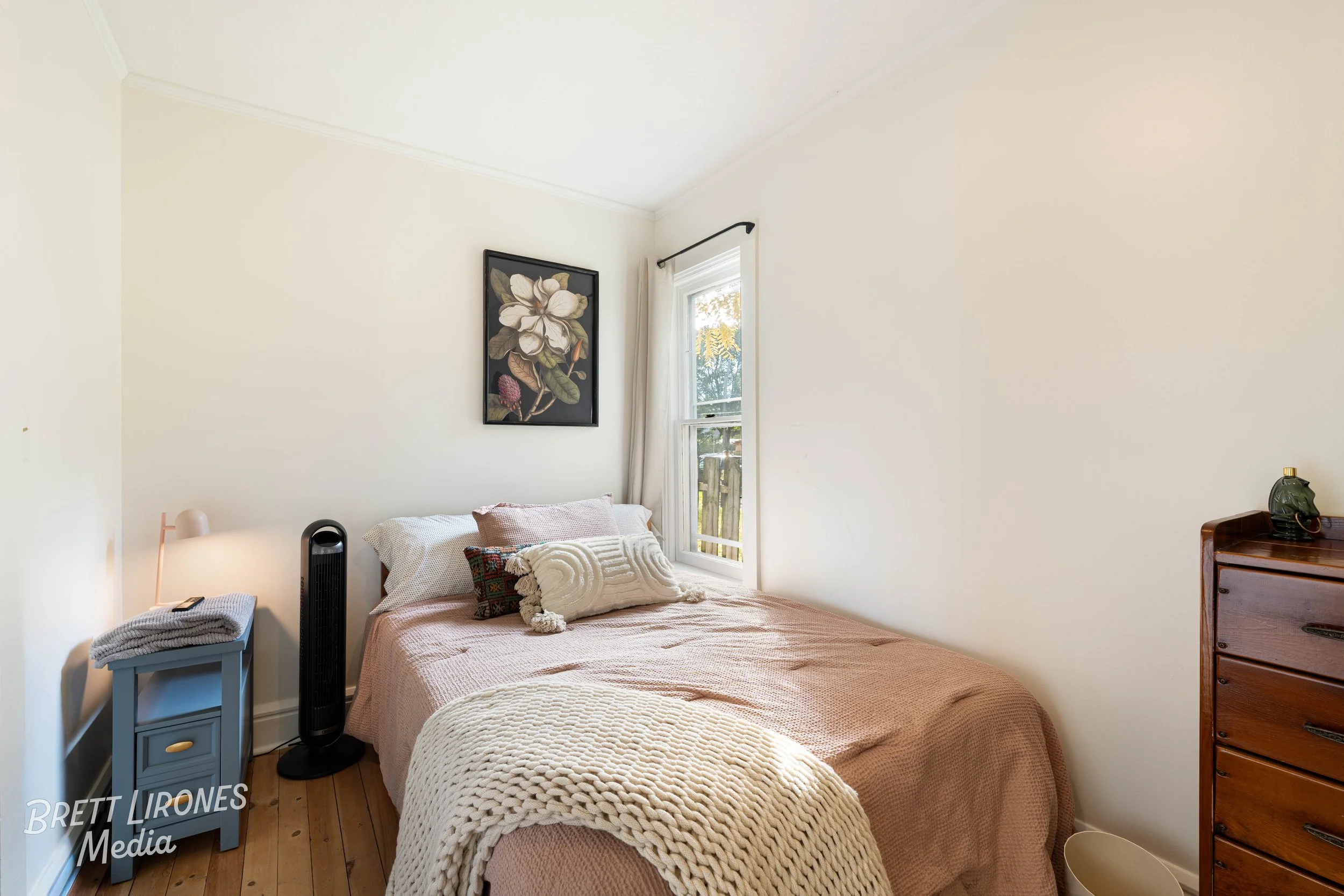 A cozy bedroom with a bed covered in a pinkish-brown blanket and decorative pillows, a small blue nightstand with a folded blanket, a tall black fan, a wooden dresser, a window with white curtains, and a large floral wall art piece.