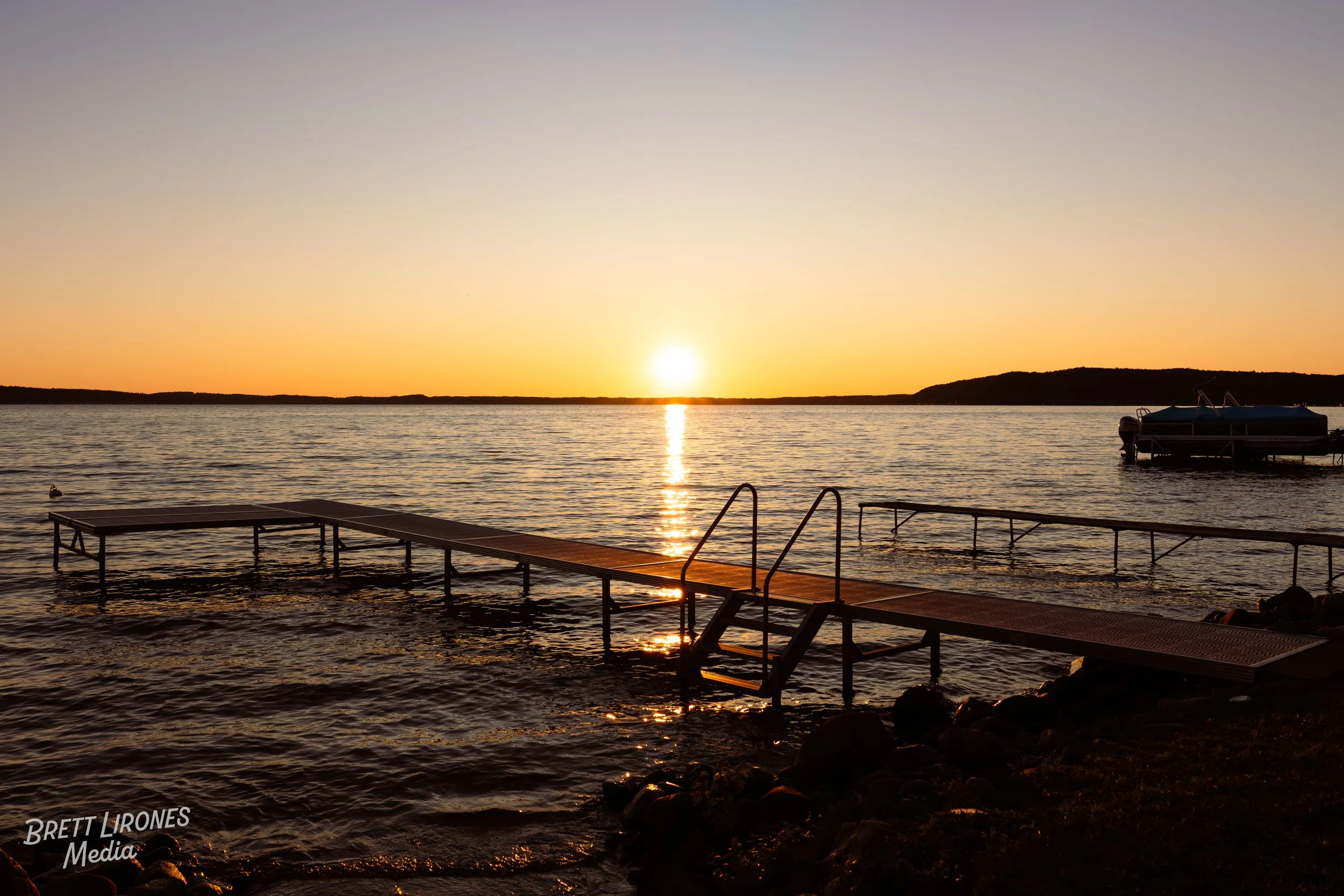 Sunset over a calm lake with a dock extending into the water and a boat anchored nearby.