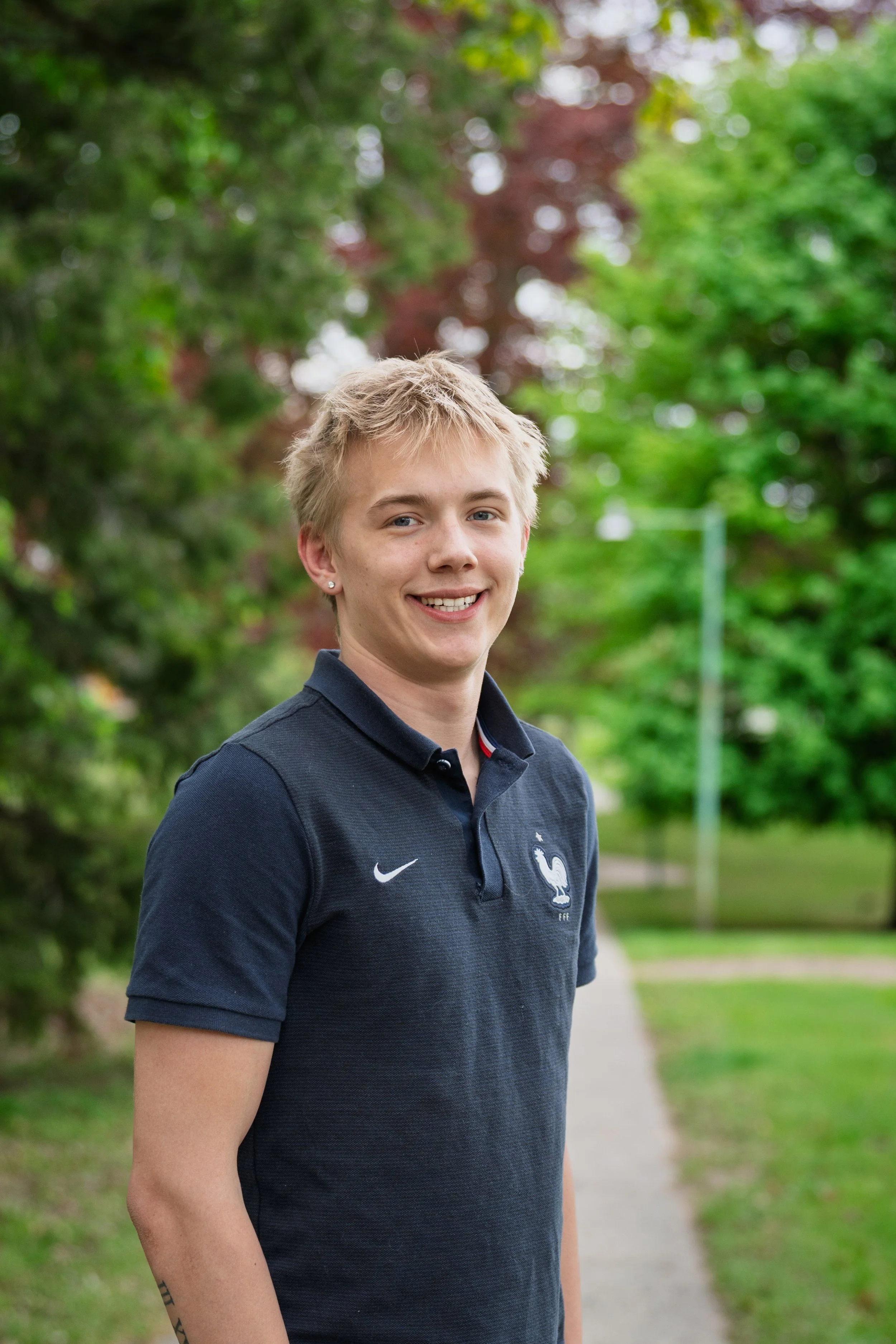 A young person with short blonde hair, smiling, wearing a navy blue shirt with the French national football team logo, standing outdoors in a park with green trees in the background.