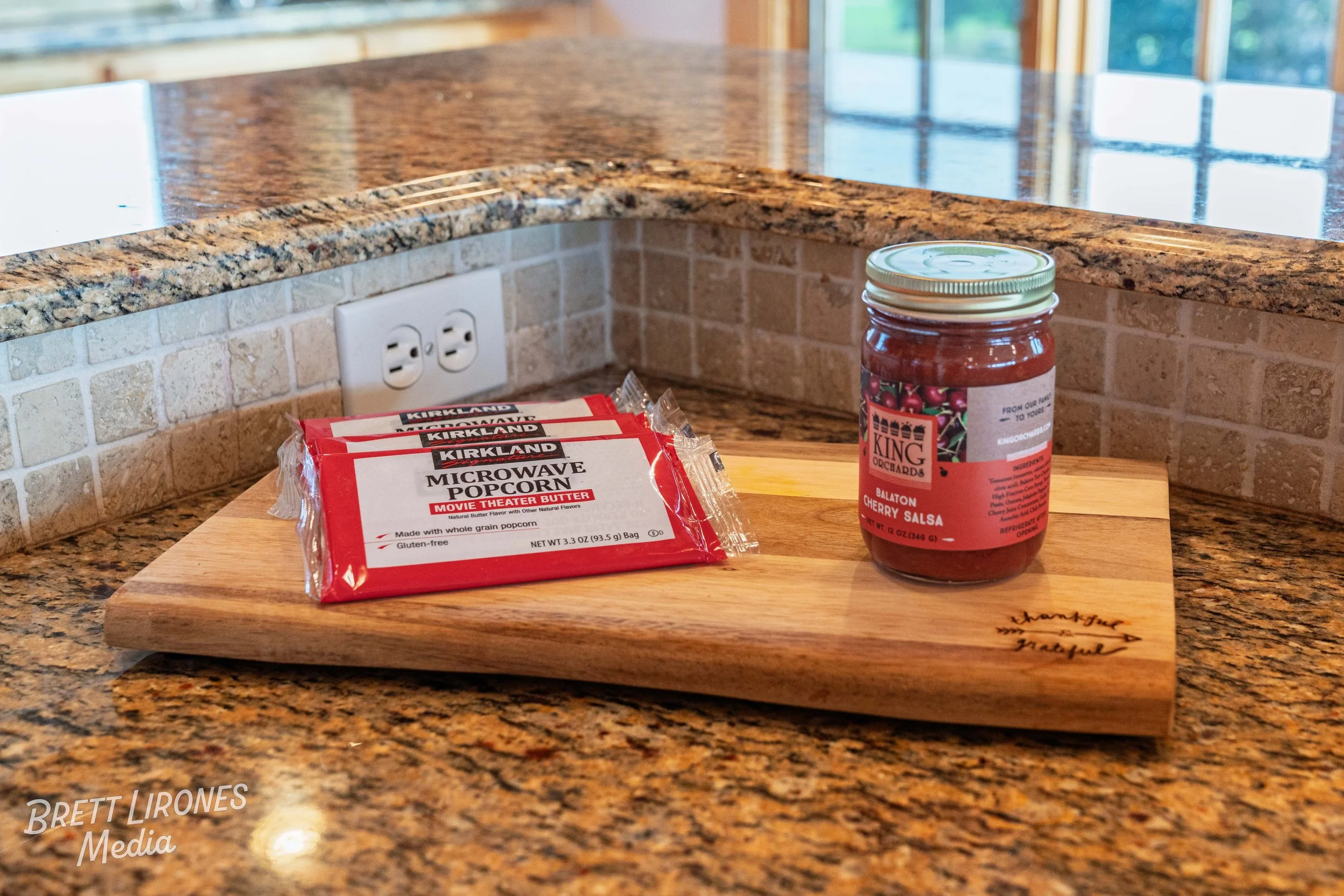 A wooden cutting board on a granite kitchen counter holds three packs of Kirkland microwave popcorn and a jar of King Orchards Balaton cherry salsa. The kitchen background includes a brick backsplash and an electrical outlet.