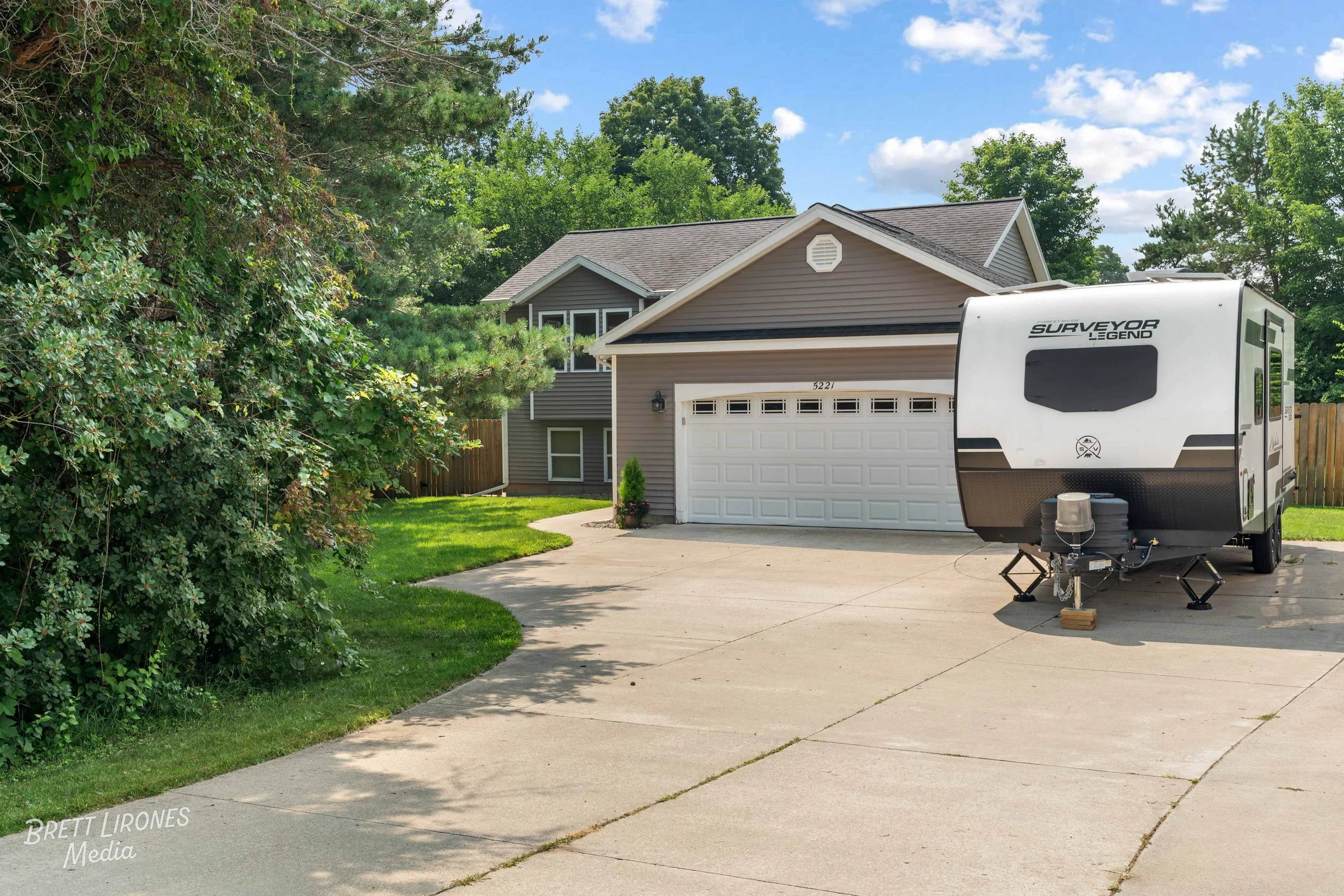 A house with a light gray exterior, a two-car garage, green lawn, and a travel trailer parked on the driveway on a sunny day with a partly cloudy sky.