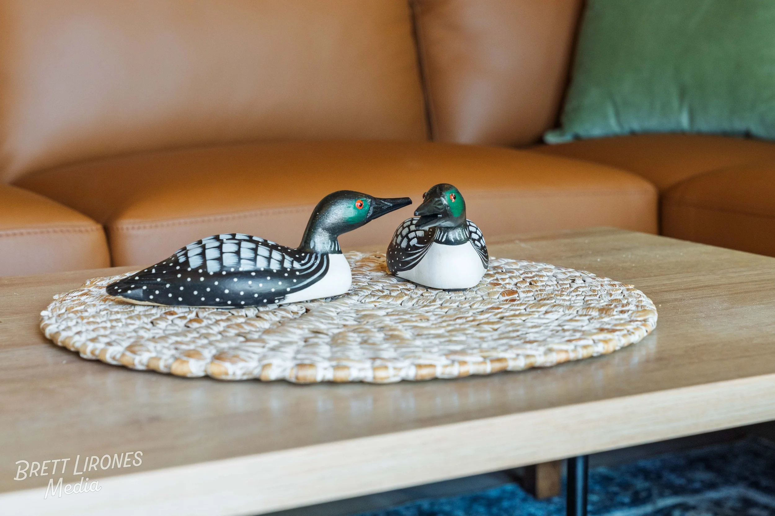 Two decorative wooden loon sculptures on a woven mat on a wooden table, with a brown sofa and a green pillow in the background.