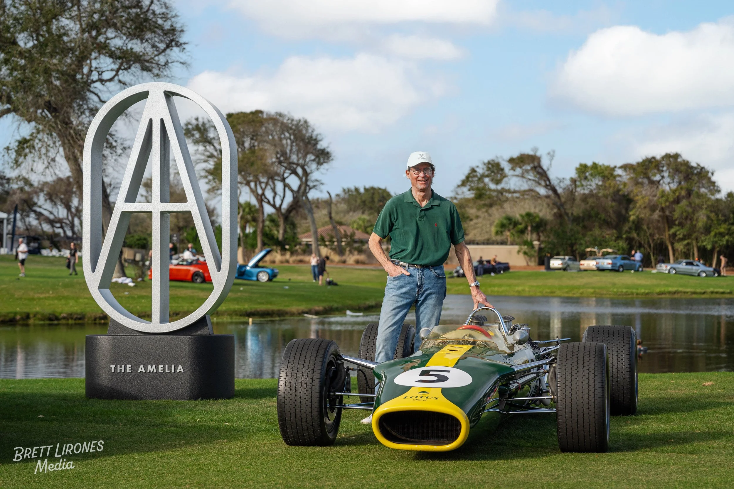 A man standing next to a vintage green and yellow Lotus racing car on a grassy area near a pond at an outdoor event, with a large modern sculpture and parked cars in the background.
