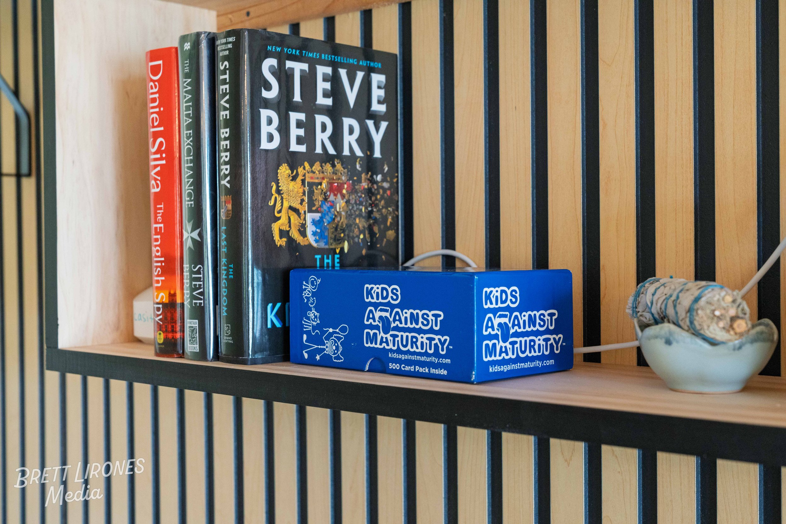 Shelf with books and game box. The books include titles by Steve Berry, Daniel Silva, and Matla Exhlange. The game box is for 'Kids Against Maturity' with 500 cards inside, placed on a black and wooden slatted shelf with shell decor on the right.