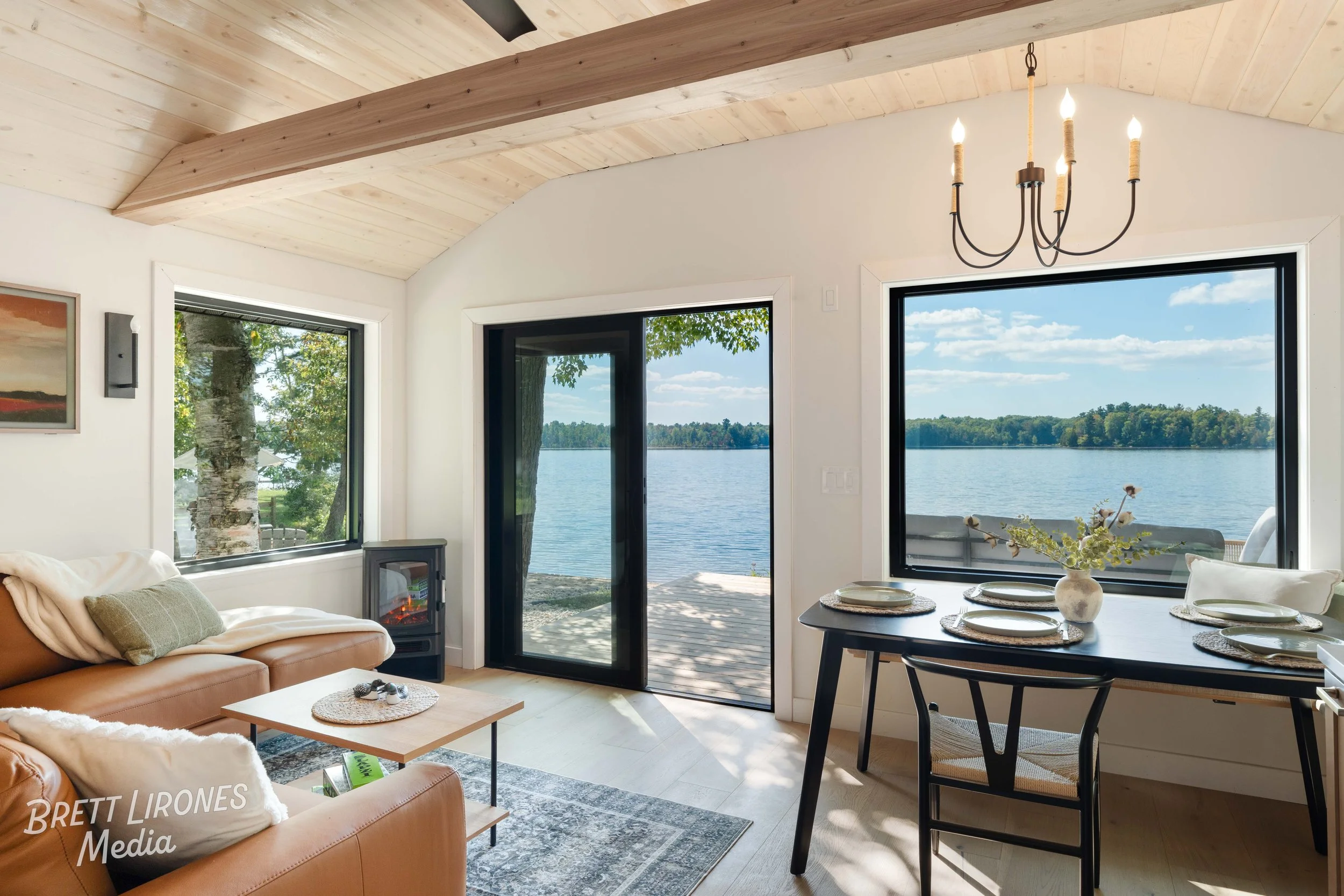 Living room with large windows and a view of a lake, featuring a leather sofa, a small coffee table, a dining table with place settings, and a chandelier, with a wooden ceiling and door leading to an outdoor deck.