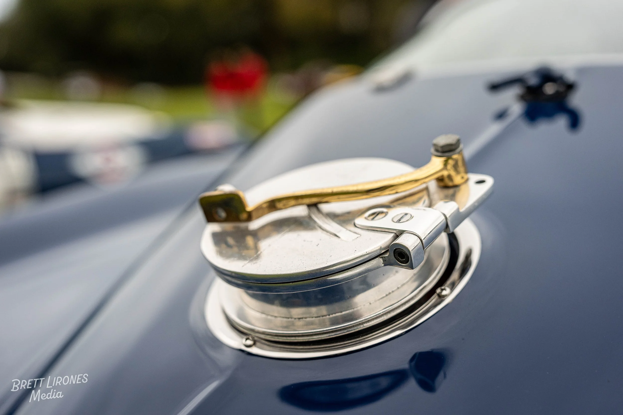 Close-up of a vintage car's fuel cap with a metallic latch and hinge, mounted on a shiny dark blue/black vehicle, with blurred background including trees and other vehicles.