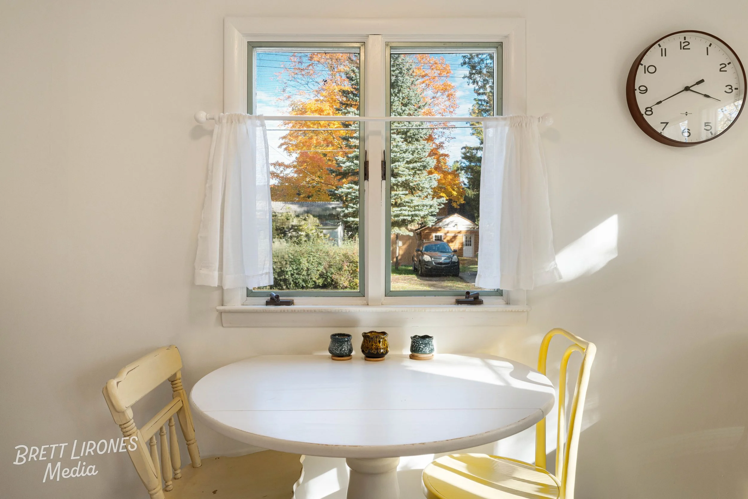Sunlit kitchen corner with a round white table, two mismatched chairs, three small decorative cups, a window with white curtains showing a view of trees and a house outside, and a wall clock showing 2:13.